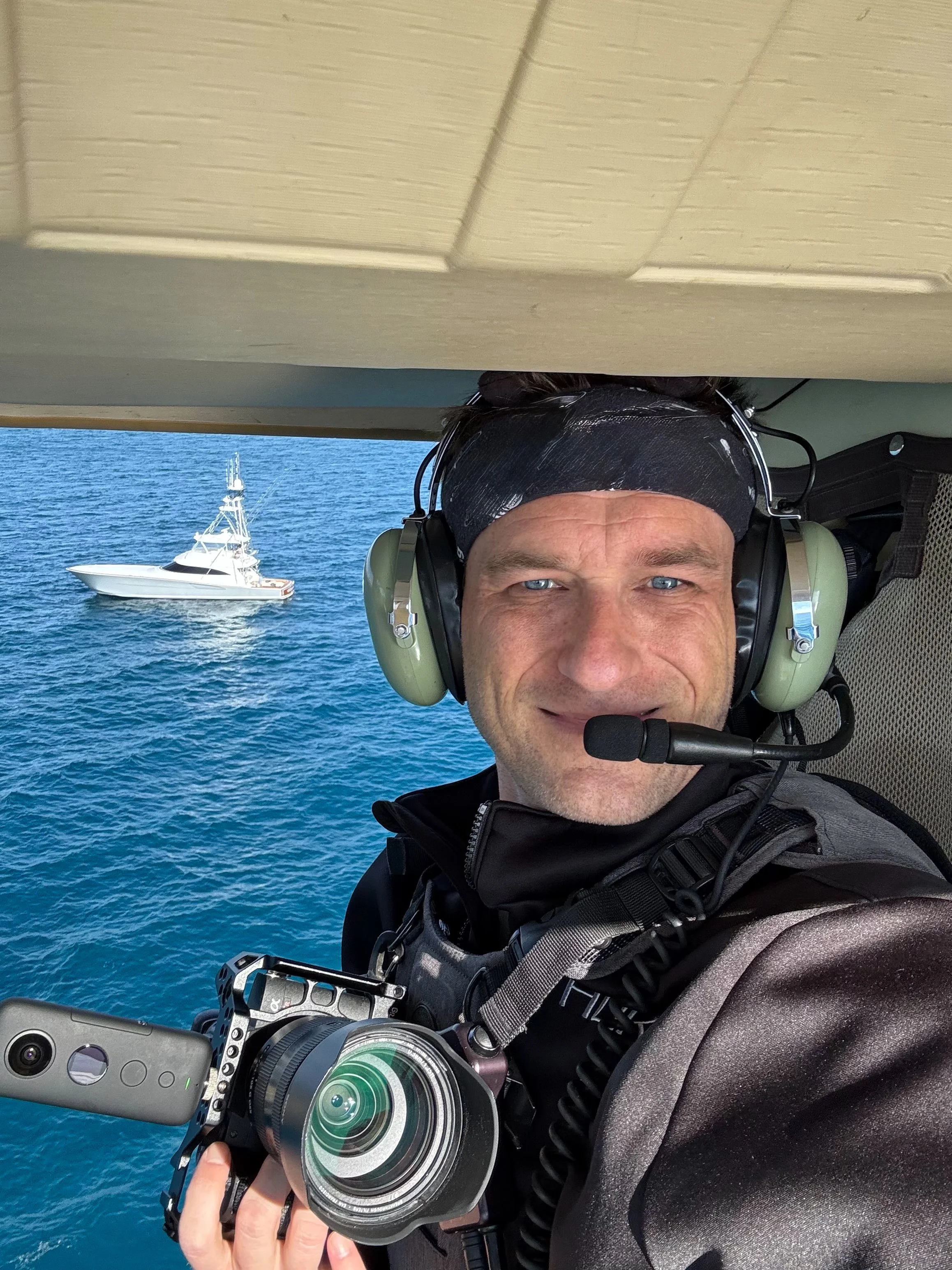 A man wearing a headset and holding a gimbal-stabilized camera taking a selfie in a helicopter over the ocean with a boat visible in the background.