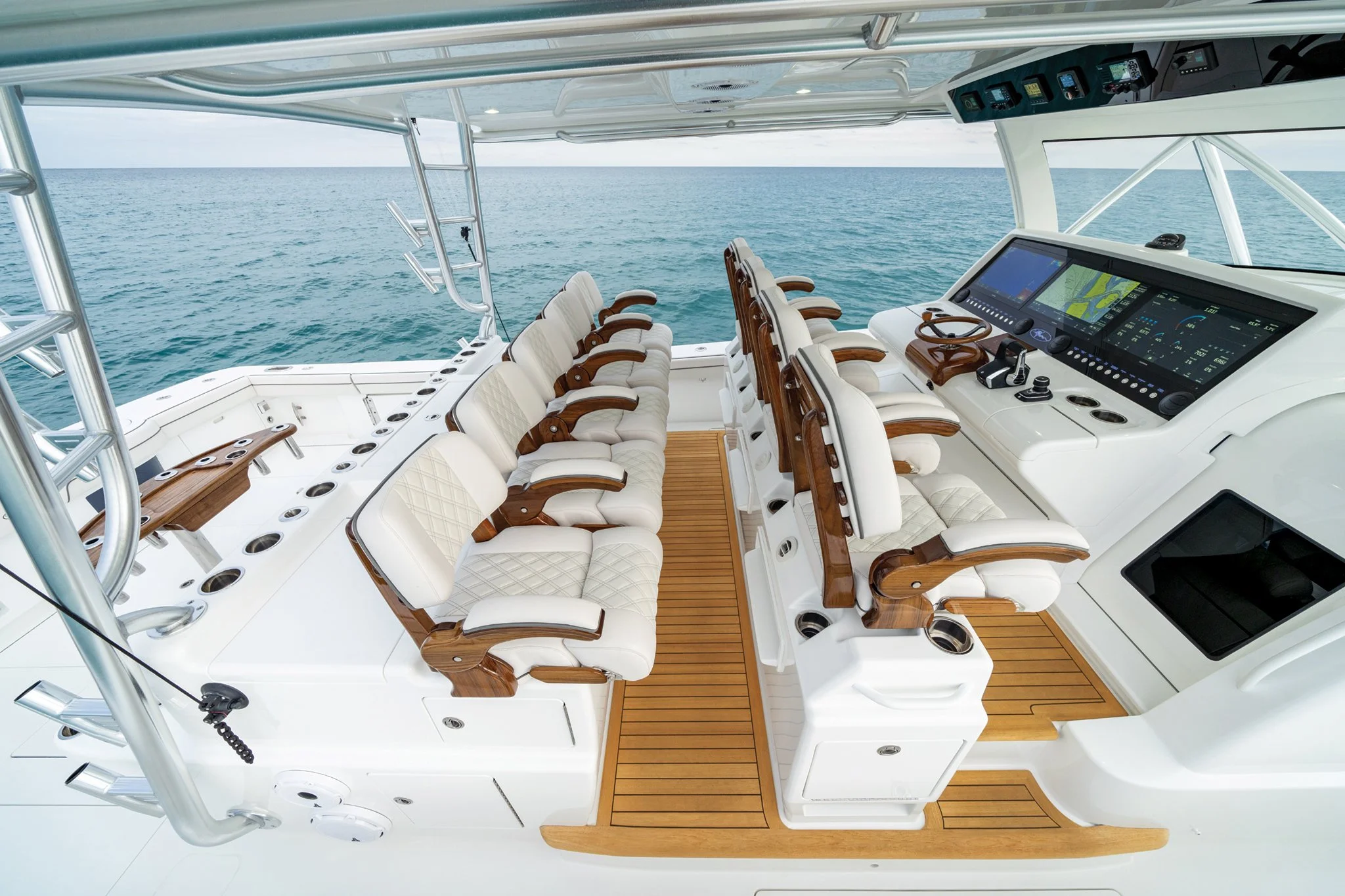 View of a boat's cockpit with multiple white leather chairs with wooden armrests, a navigation and control panel with screens, and an open view of the ocean in the background.