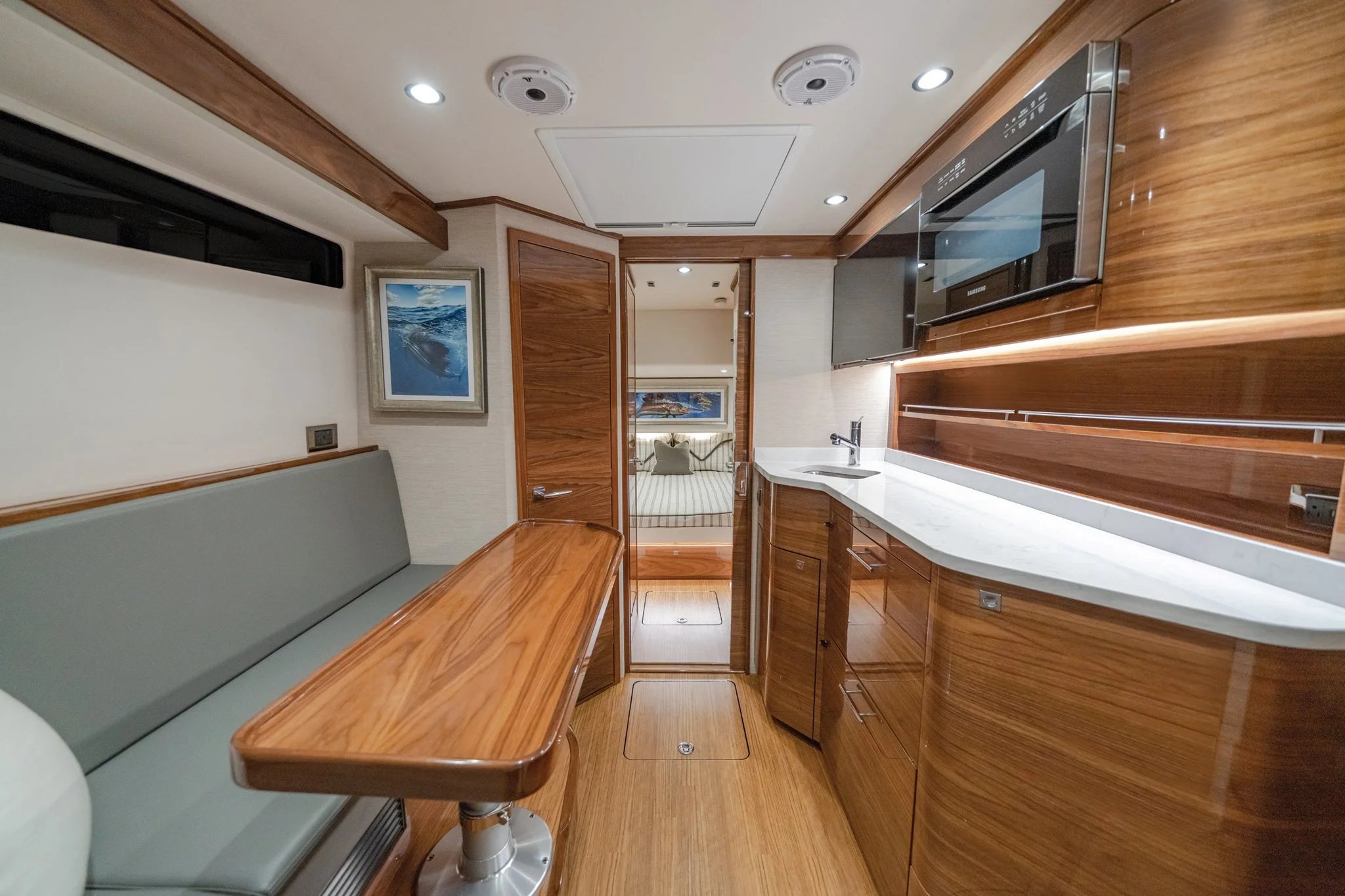 Interior of a yacht with wooden cabinetry, a small kitchen sink, a microwave, seating area, and a doorway leading to a sleeping area with a bed and pillows.
