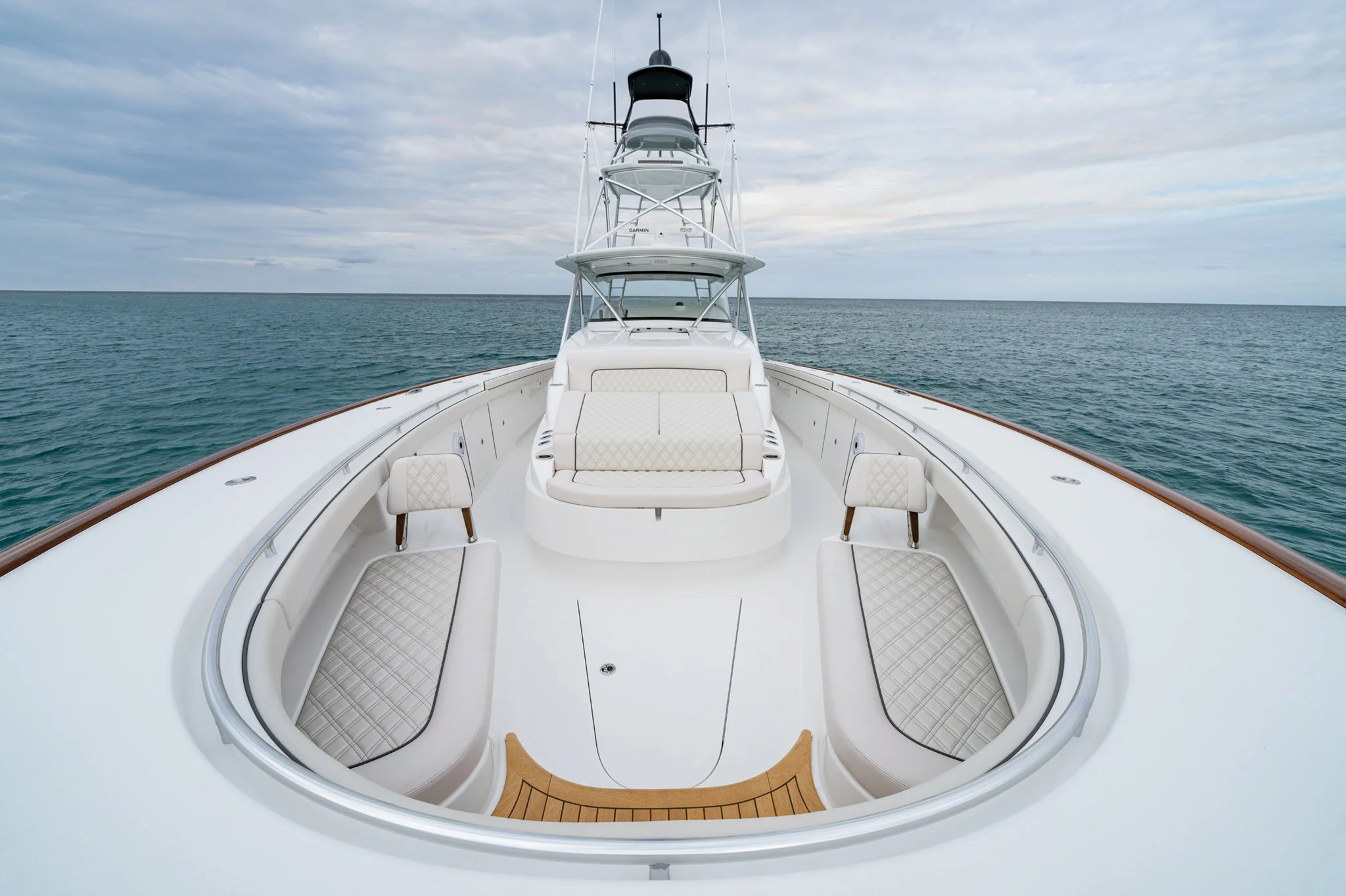 View of the front deck of a luxury white yacht with cushioned seating and two small chairs, sailing on calm ocean waters under a cloudy sky.
