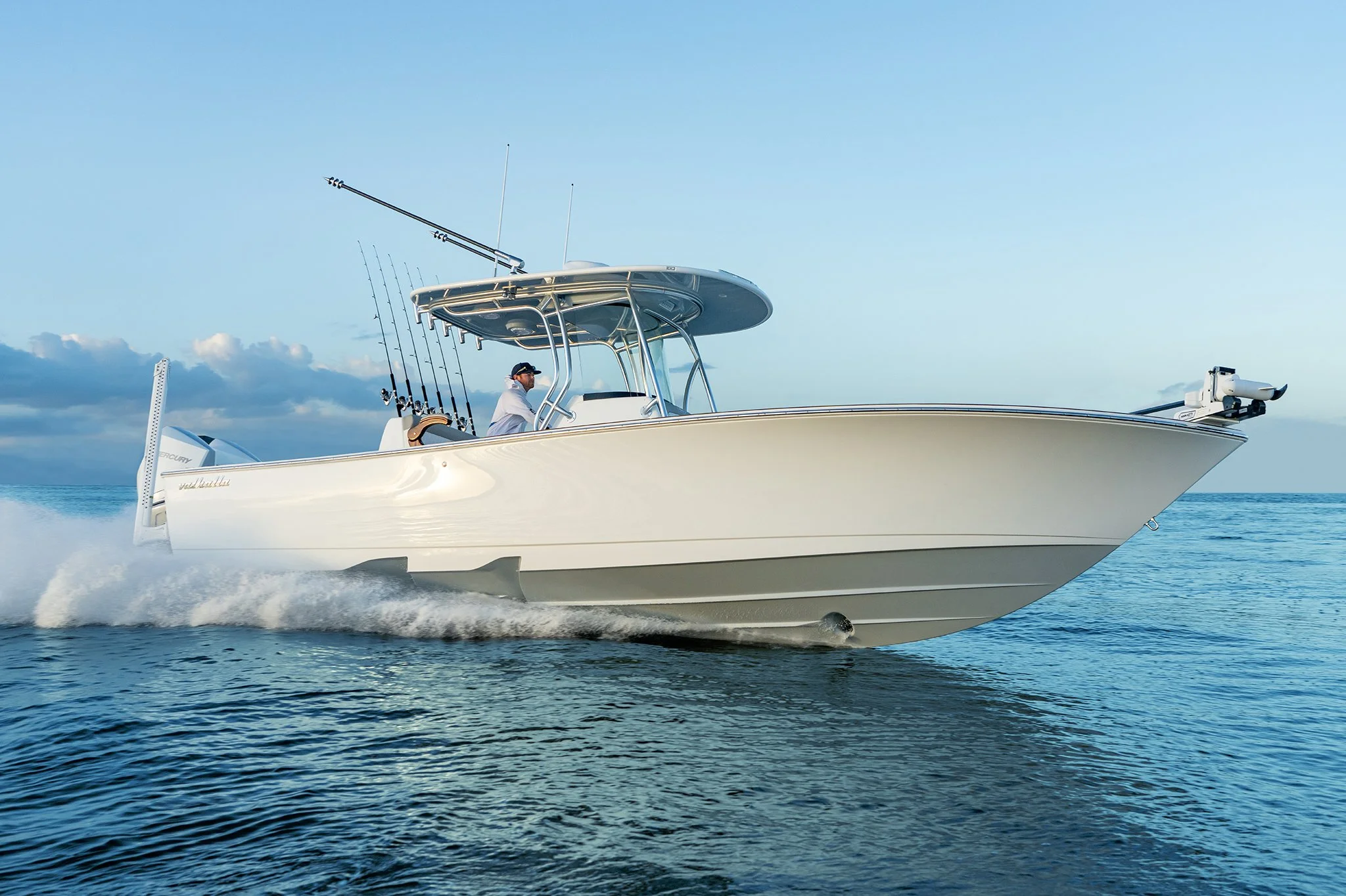 A white motorboat with a blue T-top driving on open water, with a man at the helm, two fishing rods, and a mounted trolling motor at the bow under a partly cloudy sky.