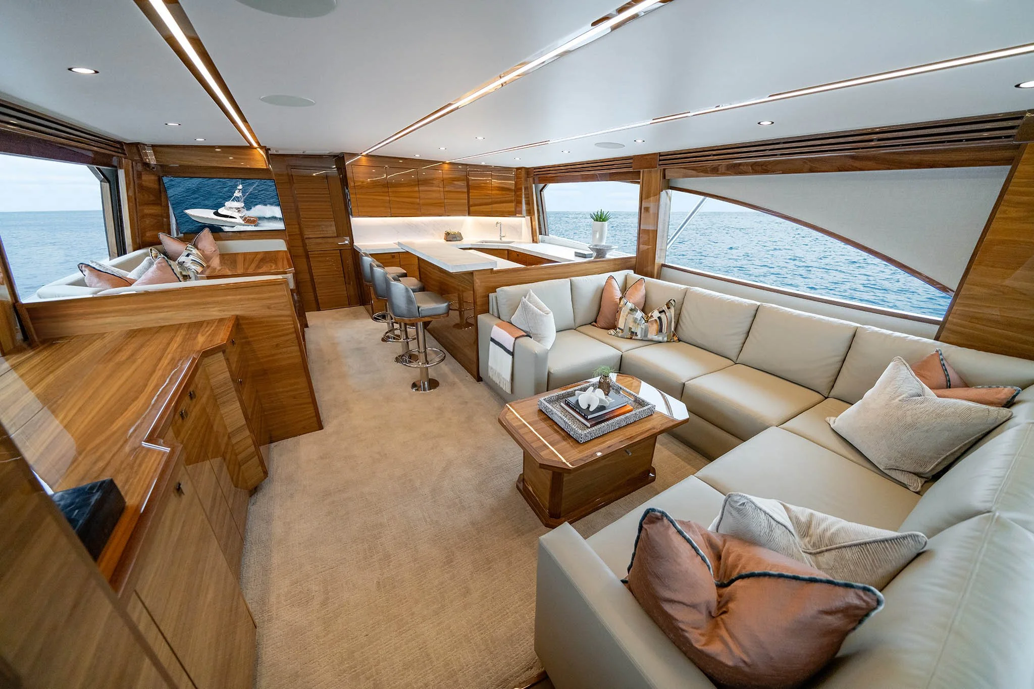 Luxury yacht interior with a cream L-shaped sofa, decorative pillows, wooden accents, barstools at a kitchen counter, a wall-mounted TV, and large windows showing the ocean outside.