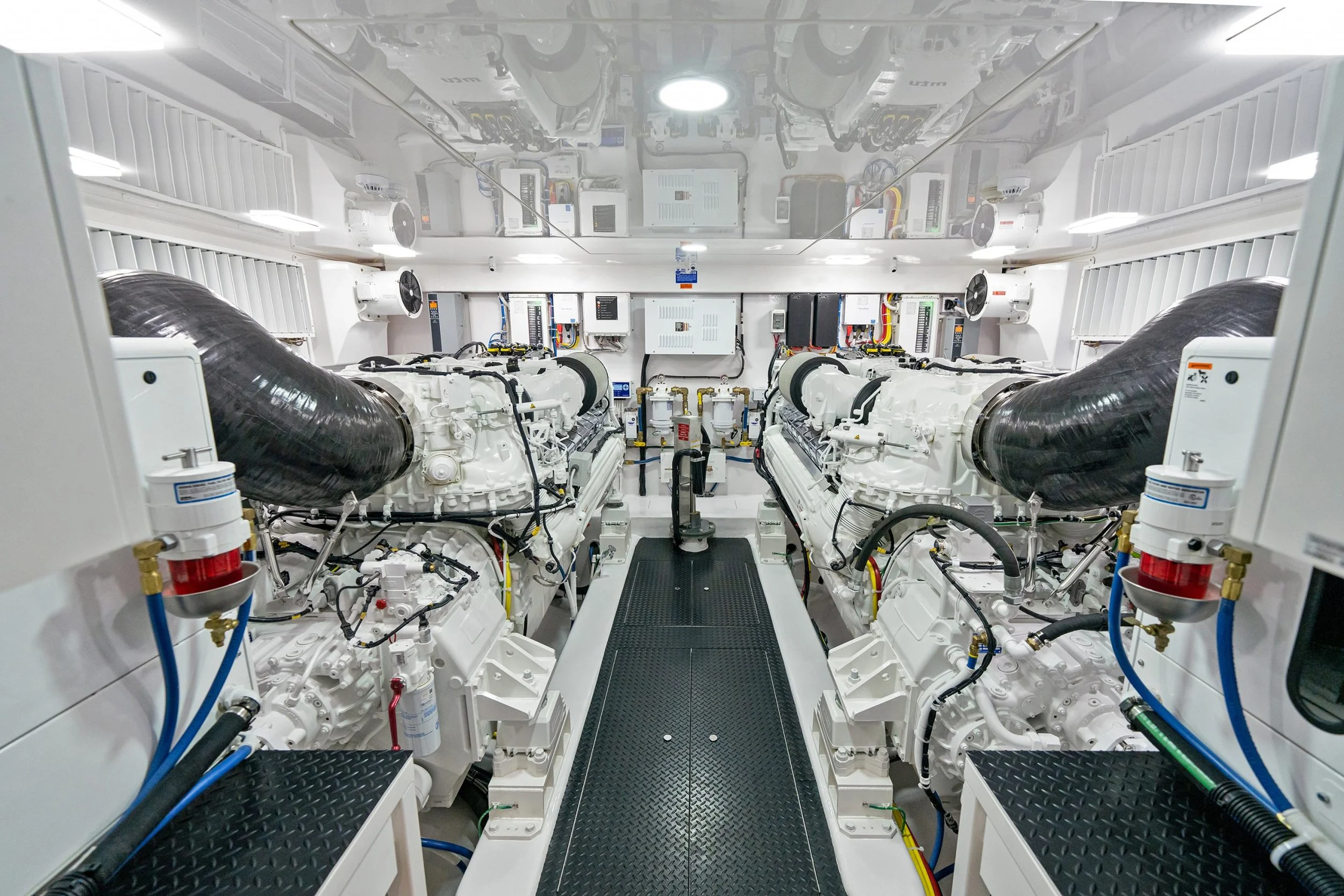 Interior of a clean, white engine room with two large diesel engines, black exhaust pipes, various control panels, pipes, and wiring, a grated walkway in the center, and bright overhead lighting.