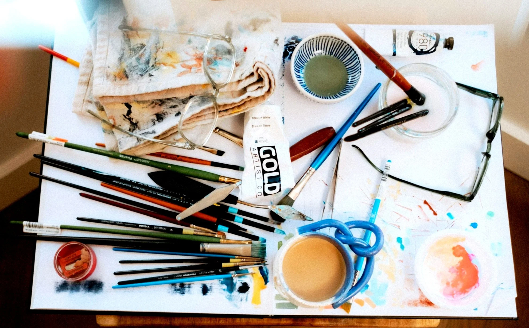 Artist's workspace with paintbrushes, paint containers, glasses, a cup of coffee, glasses, pencils, a sketchpad, and art supplies scattered on a white table.
