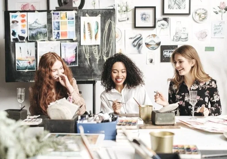 Three women sitting at a table in an art studio, laughing and enjoying a creative session. The background features a blackboard with paintings and art supplies, and sketches and artwork are displayed on the wall.