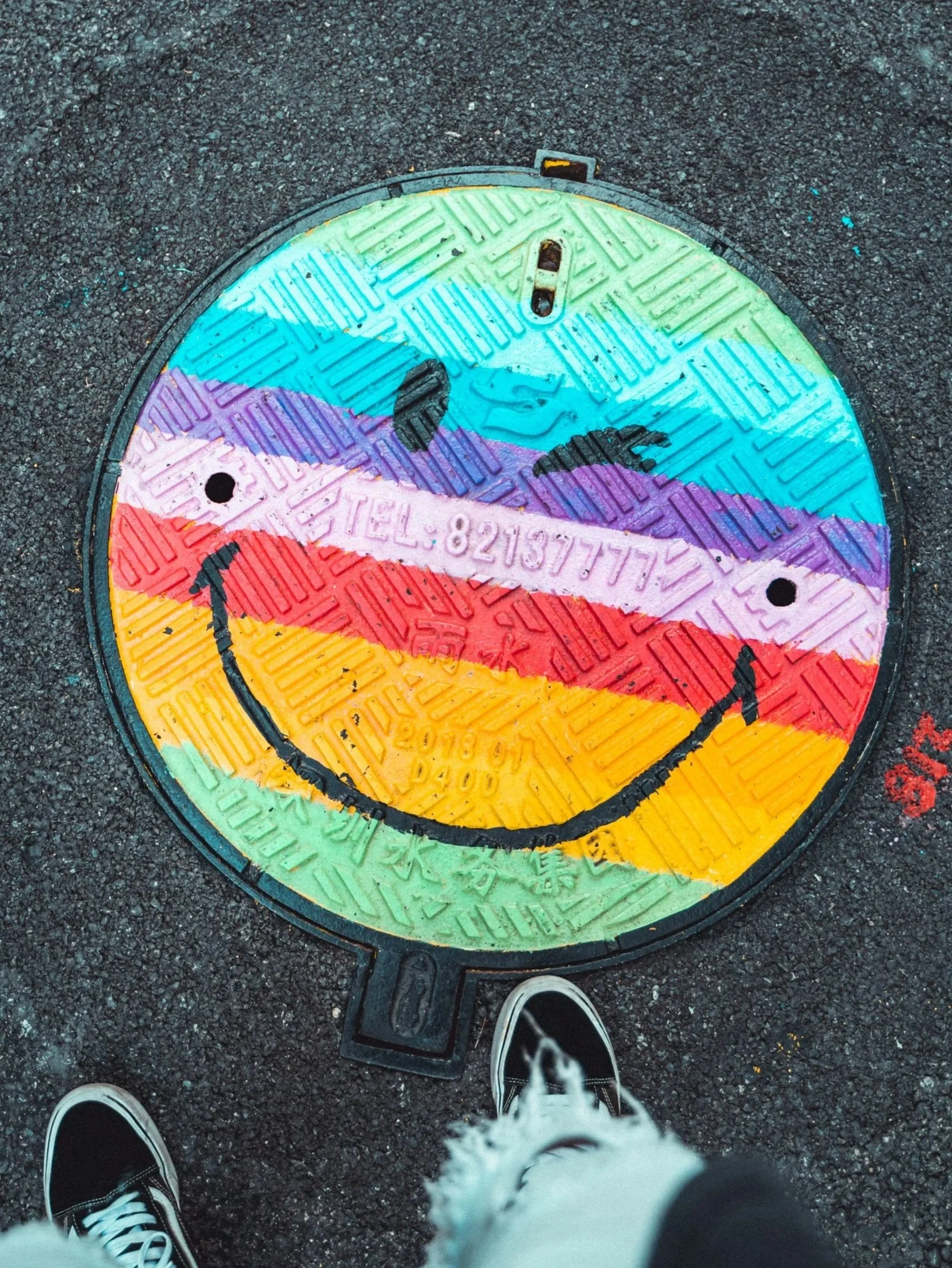 Colorful manhole cover painted with a rainbow smiley face, with the face's outline in black, on a dark asphalt street. A person's sneakers and part of their legs are visible at the bottom of the image.