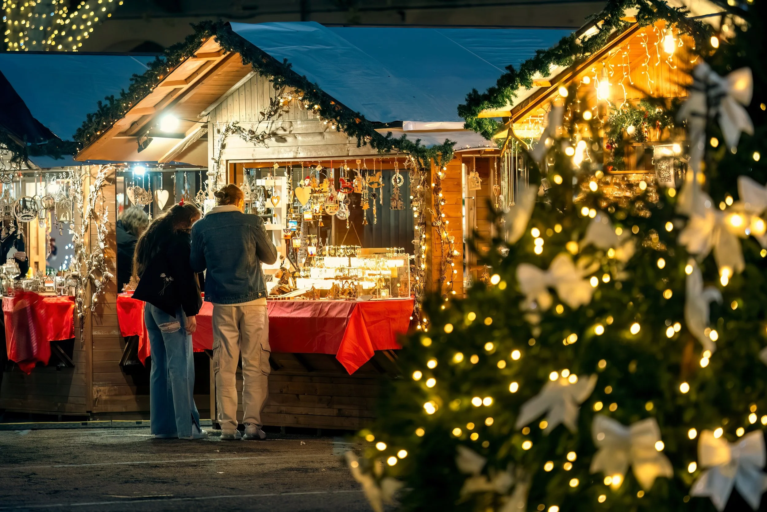 A Christmas market stall decorated with lights and gifts, with two women browsing, and a Christmas tree with white ribbons and lights in the foreground.