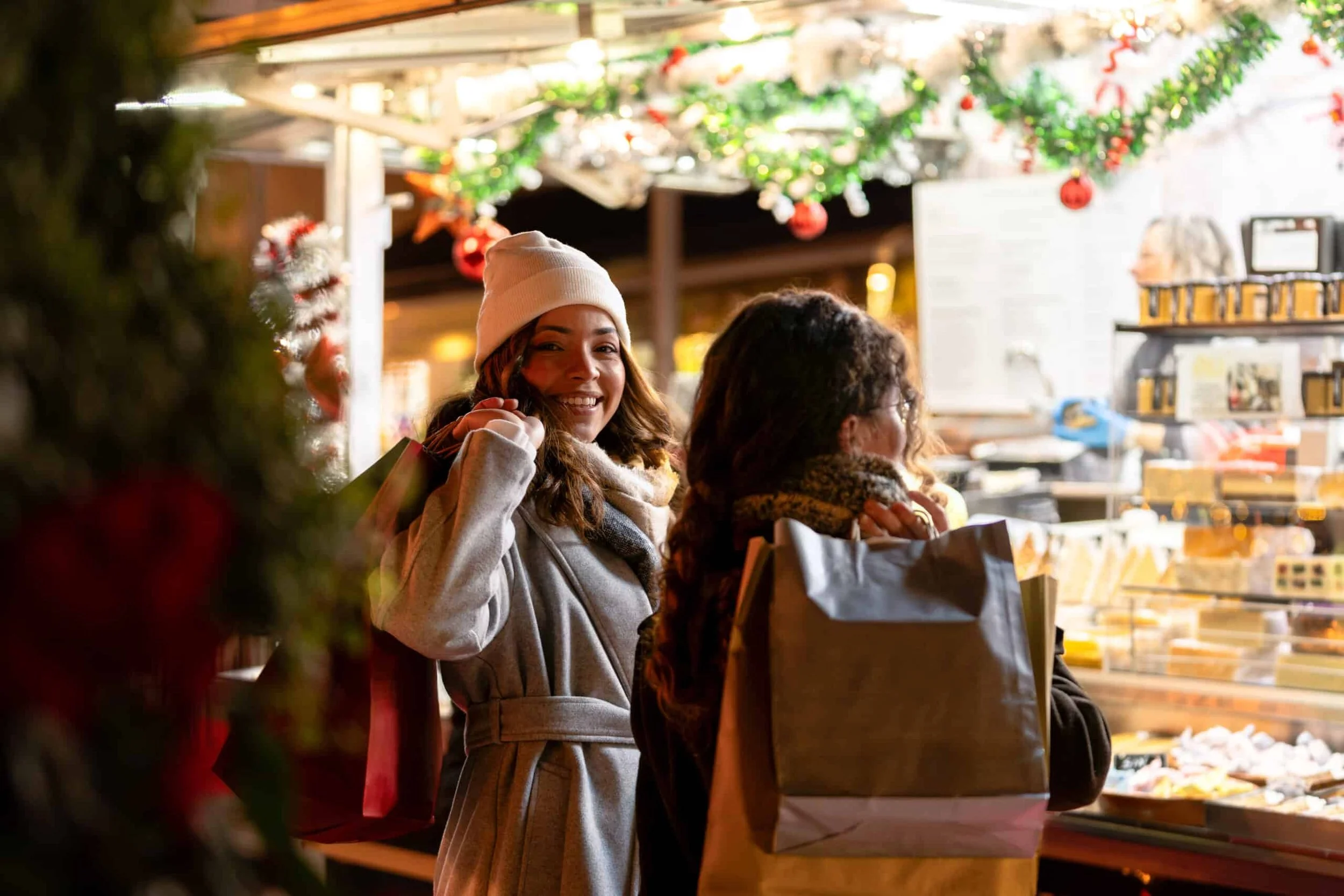 Two women shopping at a festive market stall decorated with Christmas ornaments and greenery, with one woman smiling at the camera and the other looking at items in a display case.