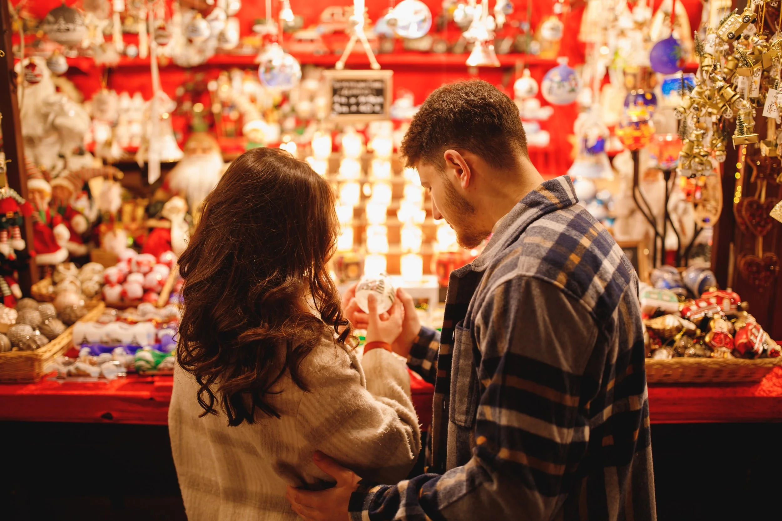 A romantic couple at a Christmas market, with decorated ornaments and holiday decorations in the background.