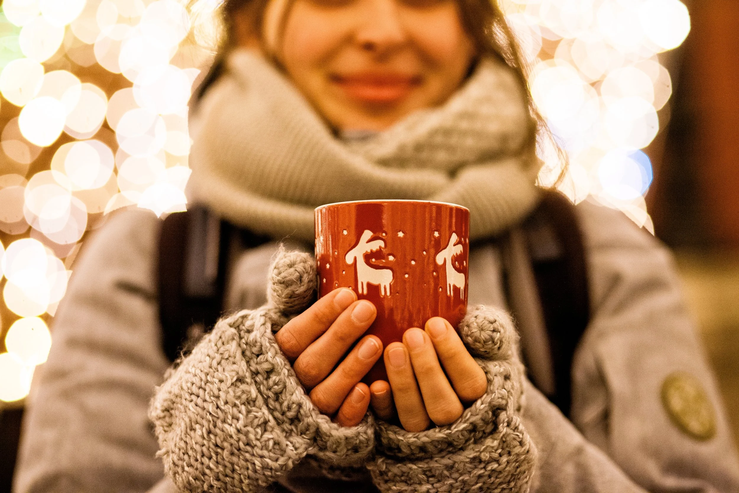 A person wearing warm gloves and a scarf holding a red mug with white reindeer designs, with blurry Christmas lights in the background.
