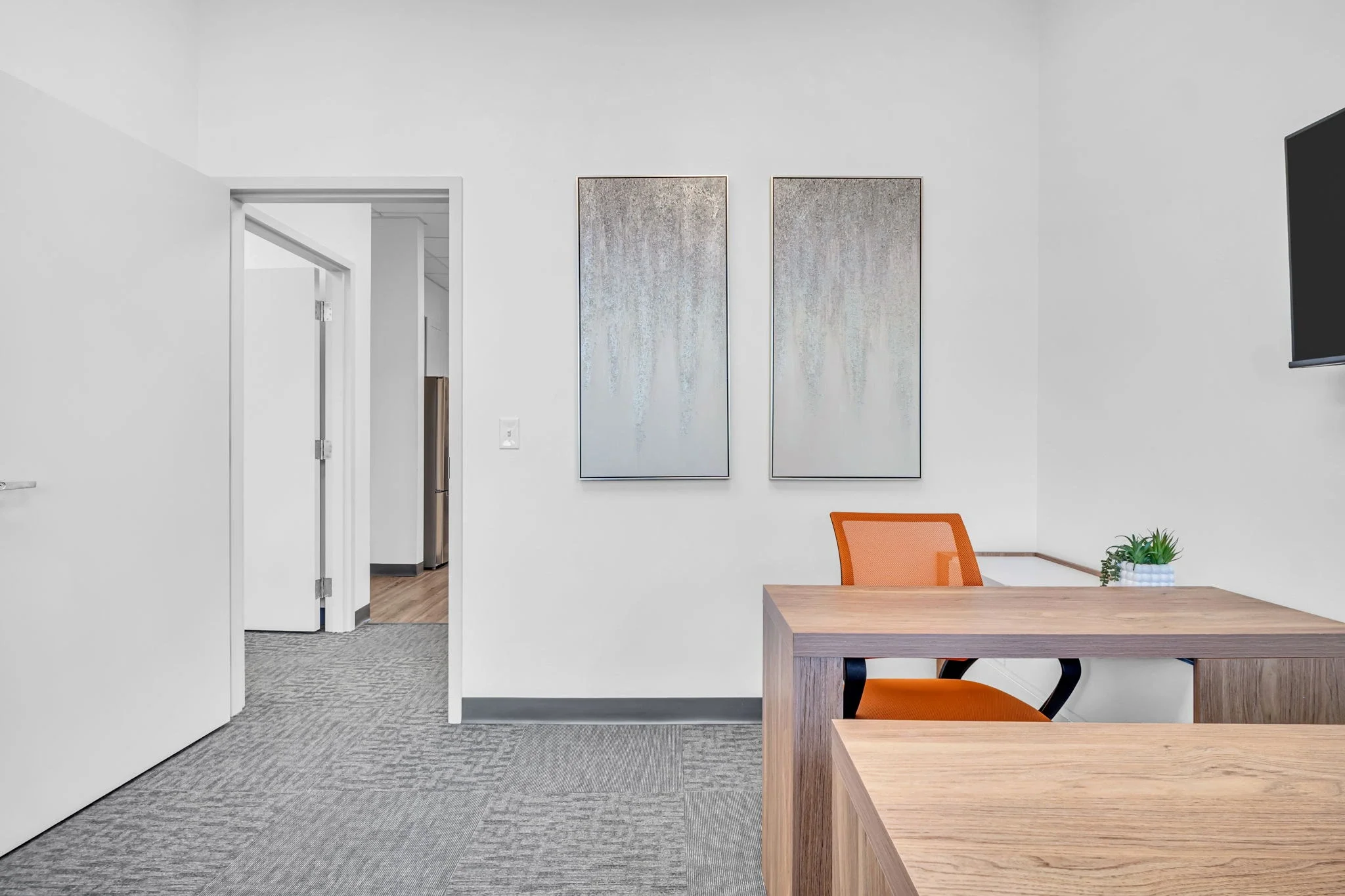 Modern office space with a wooden desk, orange chair, wall-mounted television, abstract two-panel artwork, small potted plant, and open doorway leading to a hallway with a refrigerator.
