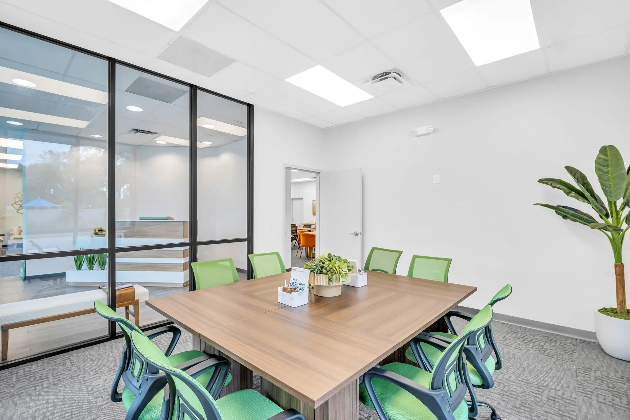 Modern conference room with a wooden table, green chairs, large potted plant, and glass wall separating an adjacent room