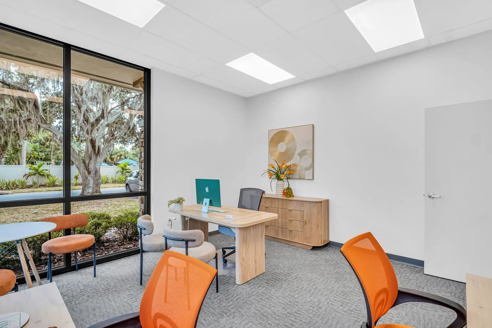 Office with wood desk, orange and beige chairs, a sideboard with a flower vase, large window with outdoor view, light-colored walls and ceiling, artwork on the wall.