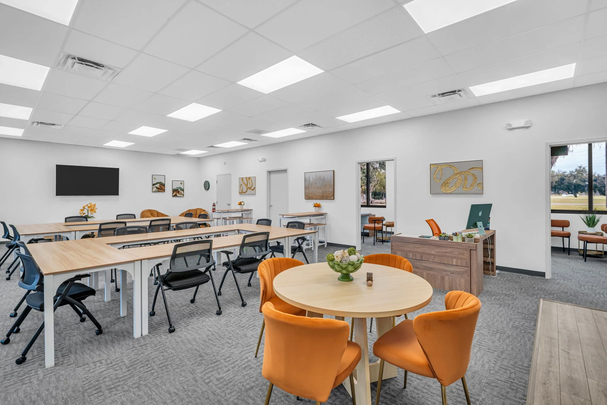 A spacious, well-lit conference or meeting room with white walls and ceiling. The room has multiple tables with black and light wood chairs, some orange chairs around a round table in the foreground, and decorative art on the walls. There is a large flat-screen TV on the wall, and windows providing natural light. The room includes a small reception desk with an Apple computer and chairs in a separate area.
