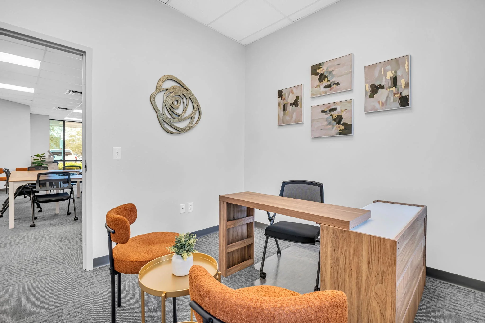 A modern office reception area with a wooden desk, two orange chairs around a small gold side table with a potted plant, artwork on the wall, and an open doorway leading to a conference room with tables and chairs.