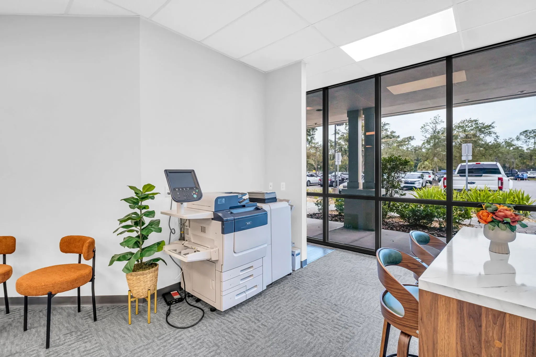 Office waiting area with orange chairs, a potted plant, a copier machine, a desk with a vase of flowers, and large windows showing a parking lot outside.