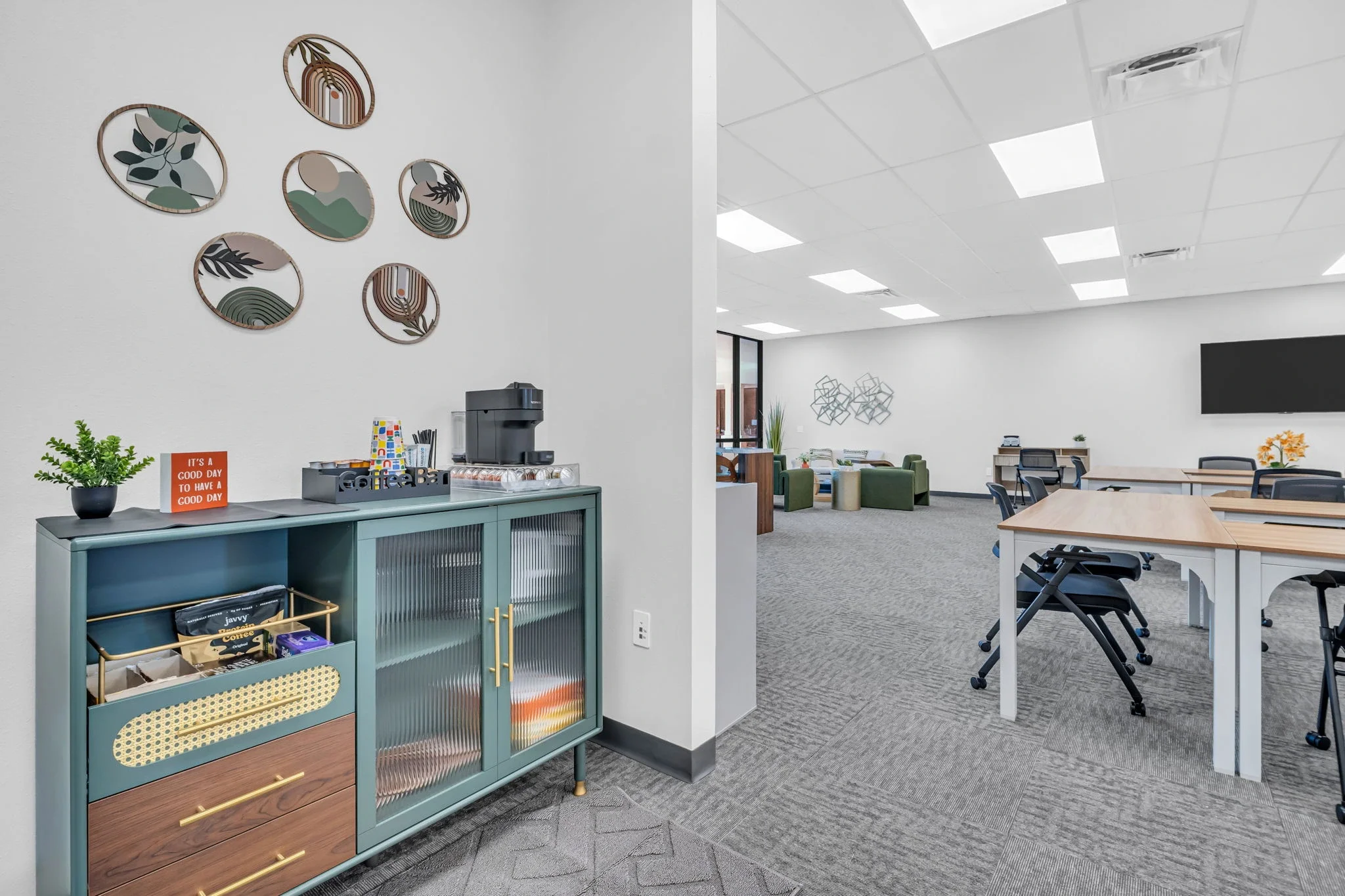 Break area with a blue cabinet holding coffee supplies, a coffee maker, and decorative wall art in an office break room. The main office space with desks, chairs, and wall decorations is visible in the background.