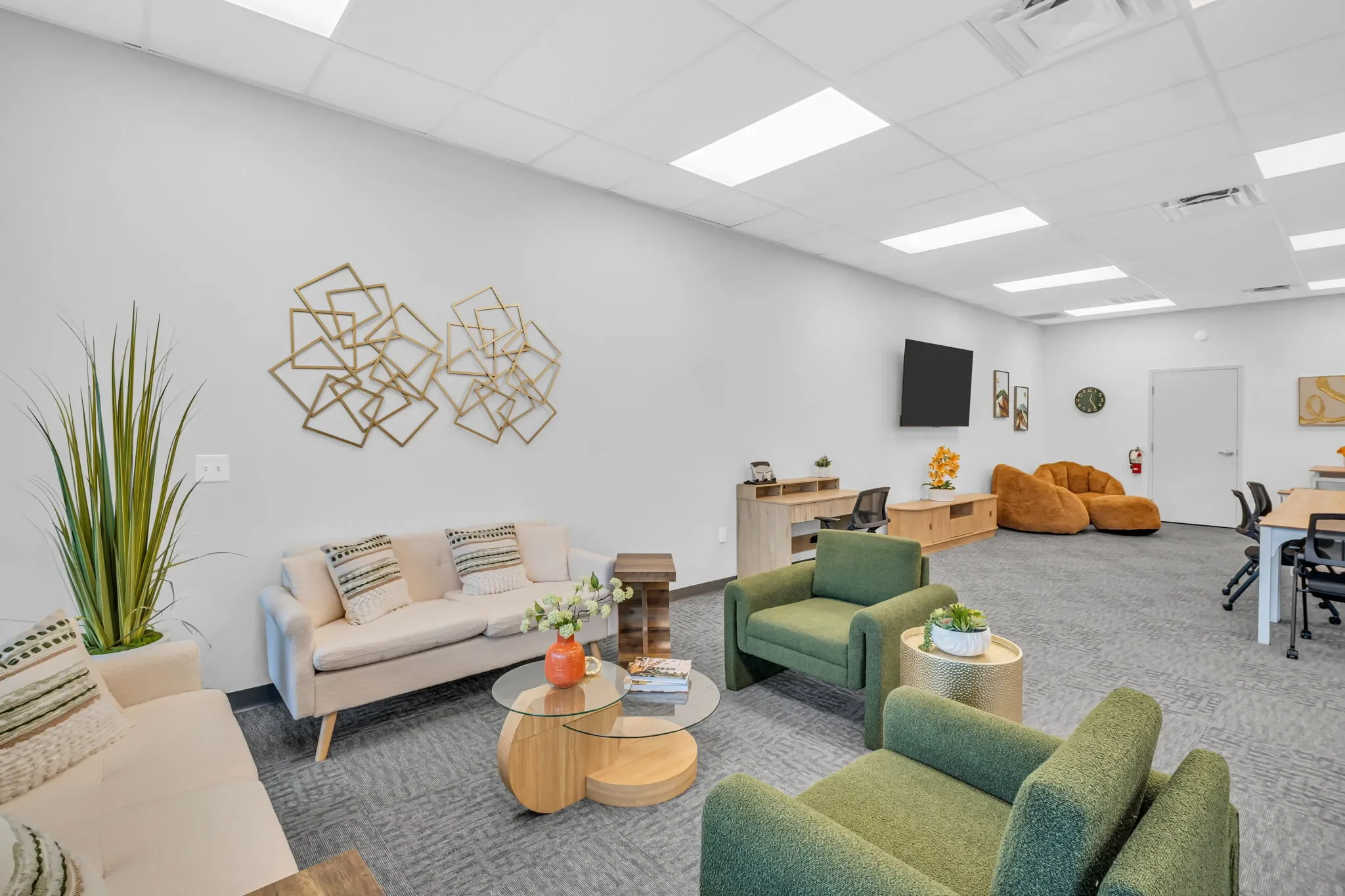 Office lounge with beige and green armchairs, white couch, decorative wall art, TV, and various furniture and decor items.