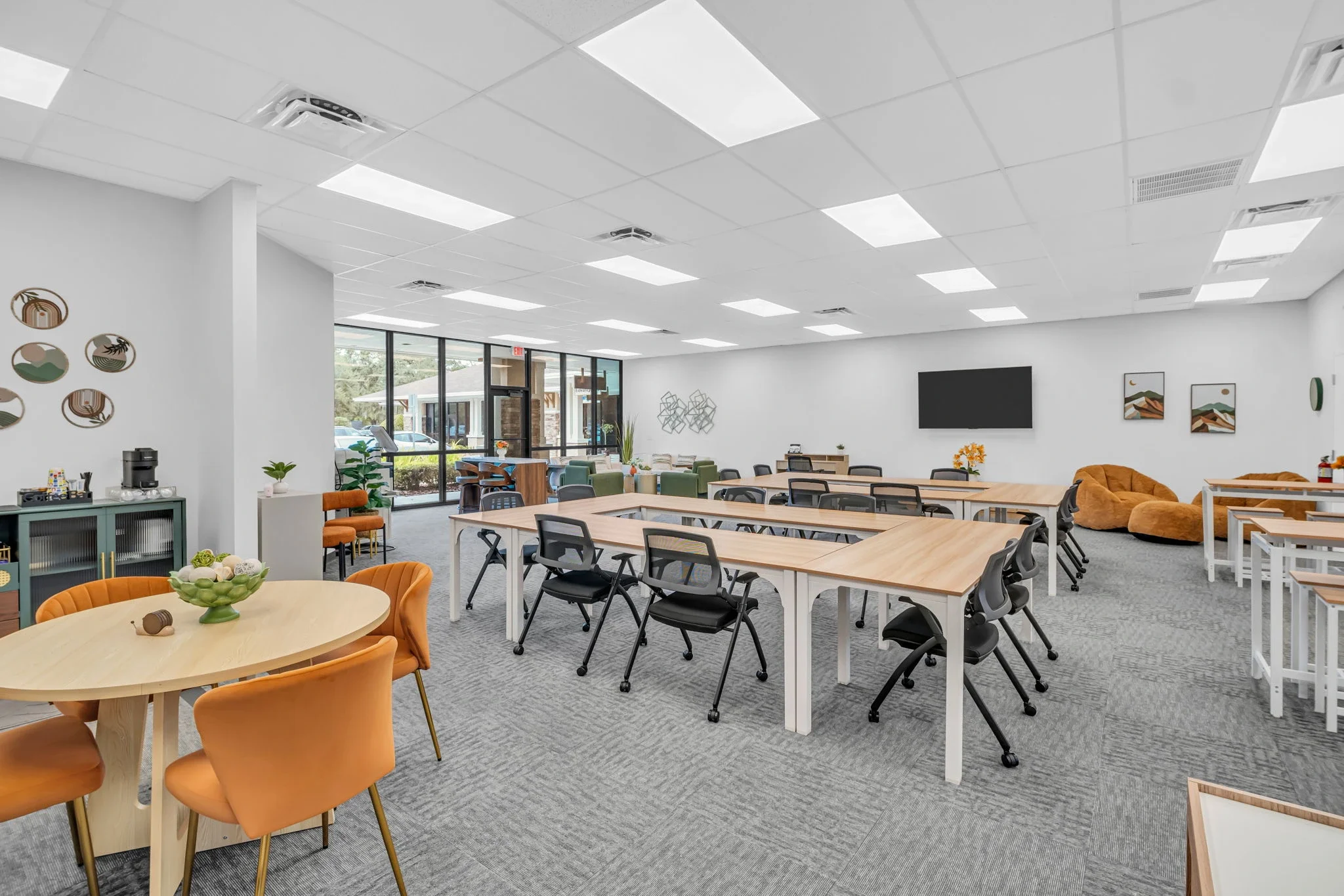 Bright, modern conference or work lounge with multiple tables, chairs, bean bags, artwork, plants, and a large wall-mounted TV.