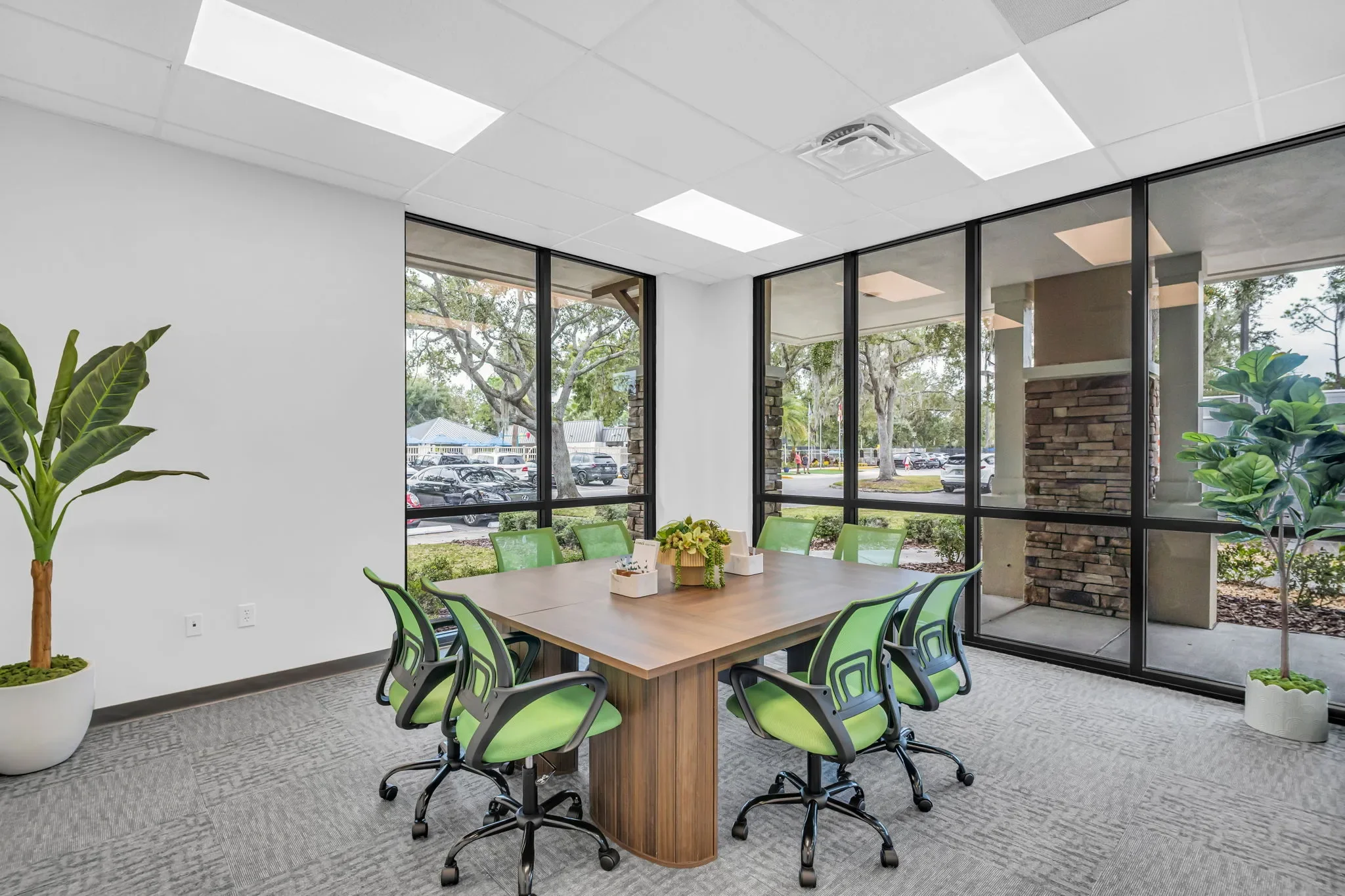 Empty office meeting room with a large wooden table, ten green office chairs, indoor plants, large window views of parked cars and trees outside, white walls, and ceiling lights.
