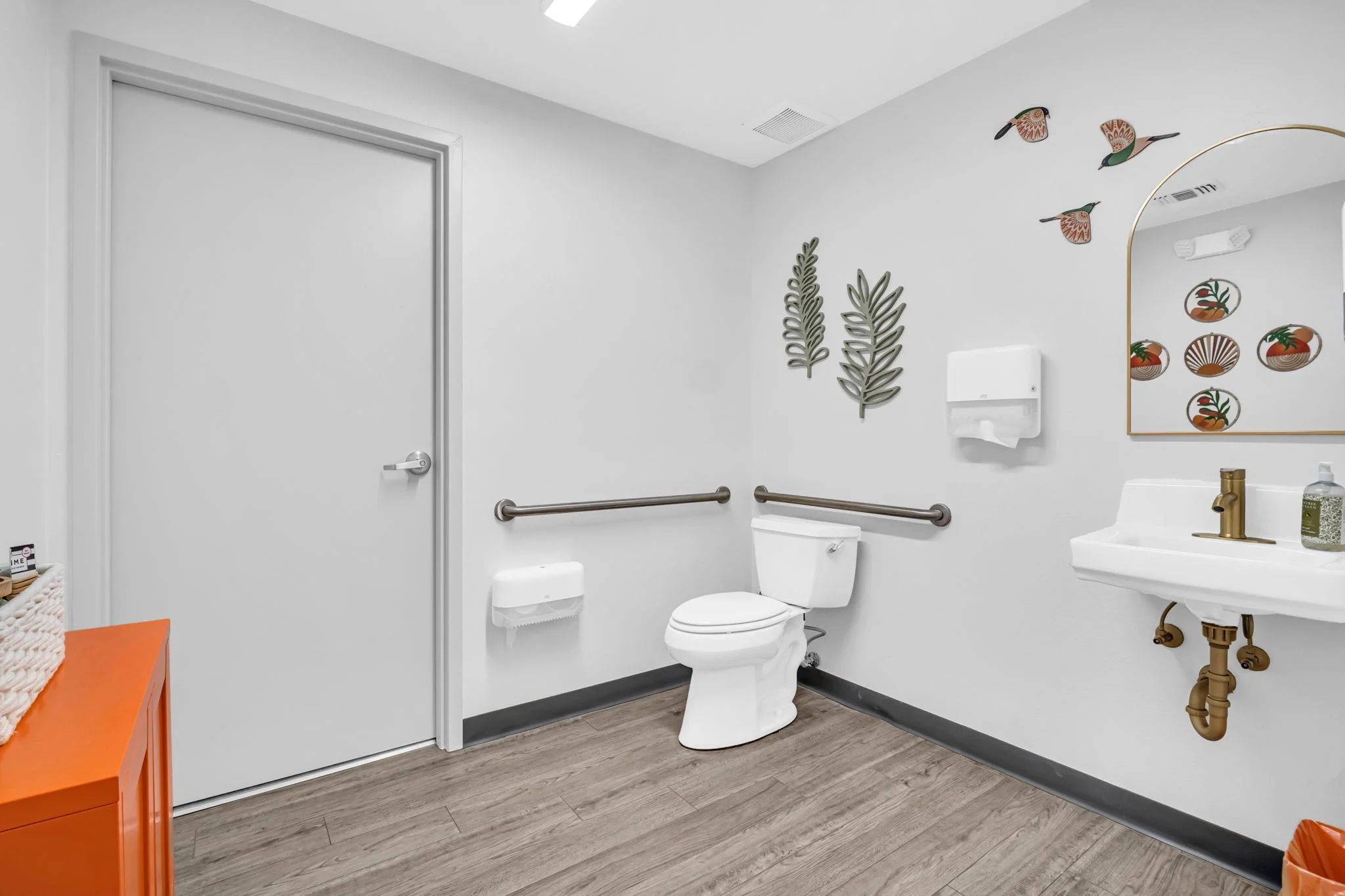 A clean, accessible restroom with a white toilet, wall-mounted paper towel dispenser, and small sink with a brass faucet. Decor includes metal leaf wall art and framed bird and plant artwork. The room features wood flooring and white walls.