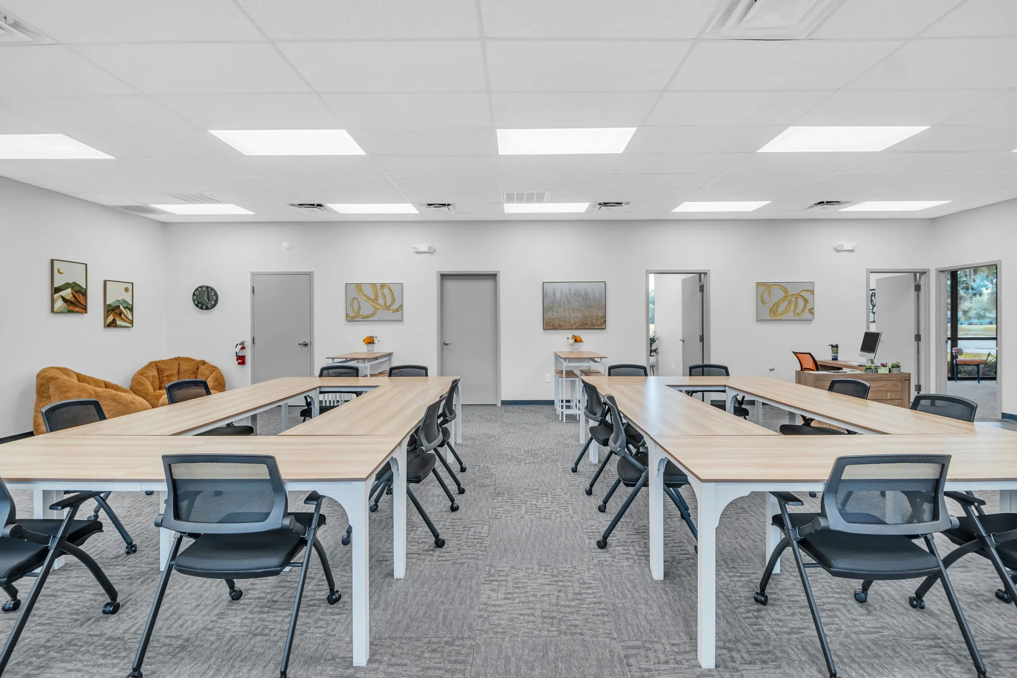Empty conference room with U-shaped desks, black chairs, artwork on white walls, and large windows.