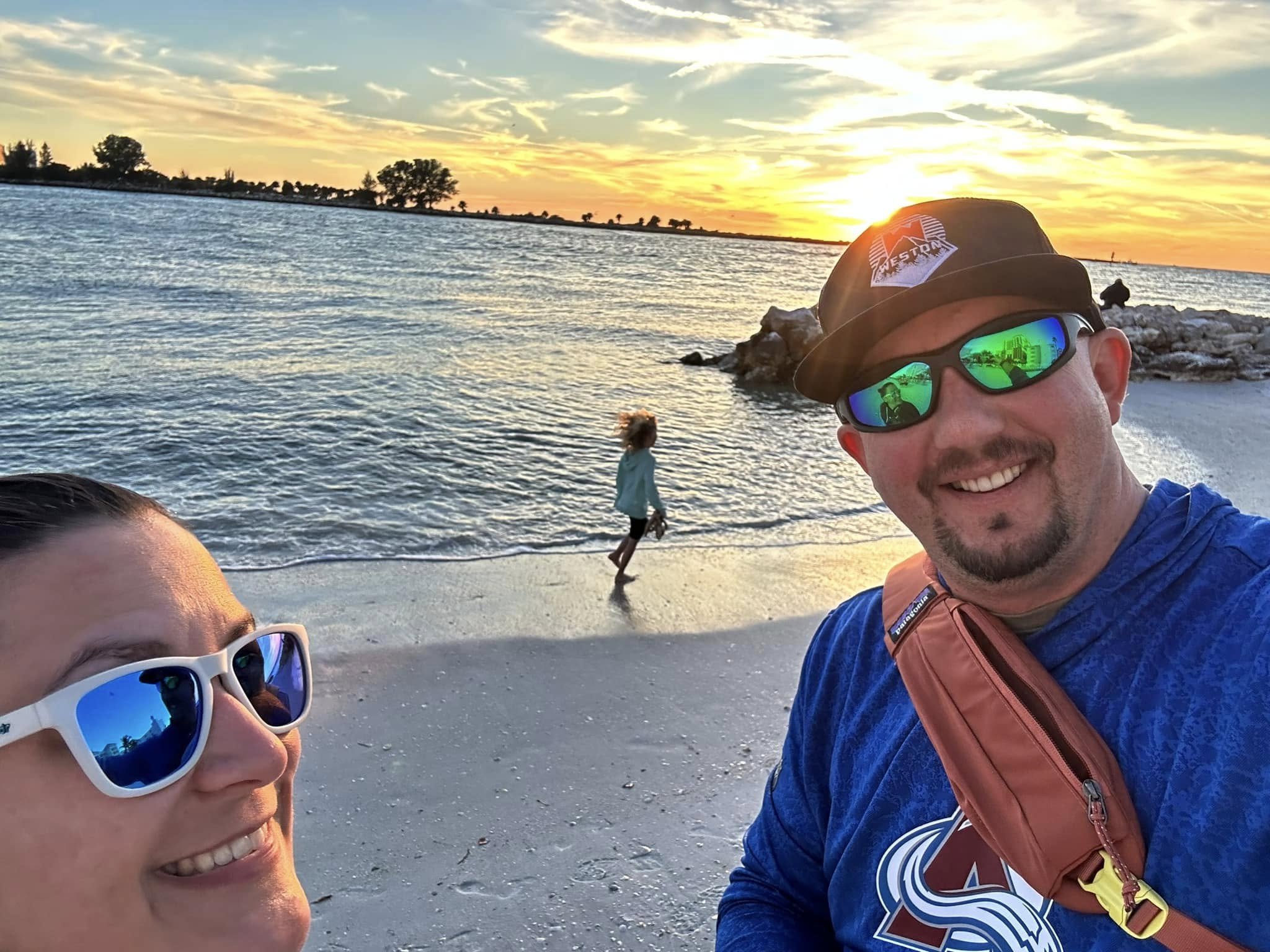 A smiling couple takes a selfie on a beach at sunset, with a child playing in the water behind them. The man wears sunglasses, a hat, and a blue jacket, while the woman wears sunglasses. The sky is colorful with the sun setting, and the ocean is calm.
