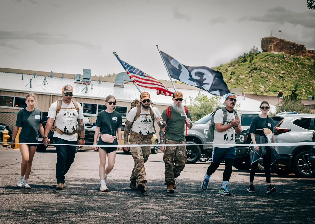Group of six young adults walking outside, four men and two women, some carrying flags and backpacks, in a parking lot near a building with a mountainous background.