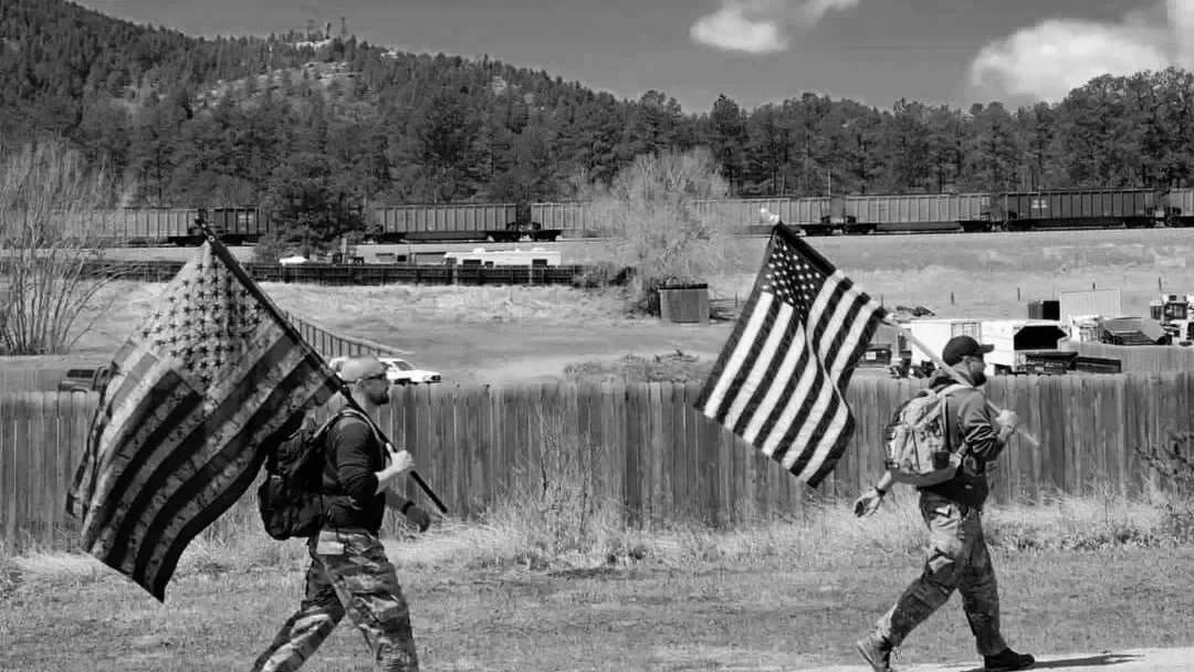 Two men walking outdoors each holding an American flag, with train tracks and trains in the background.