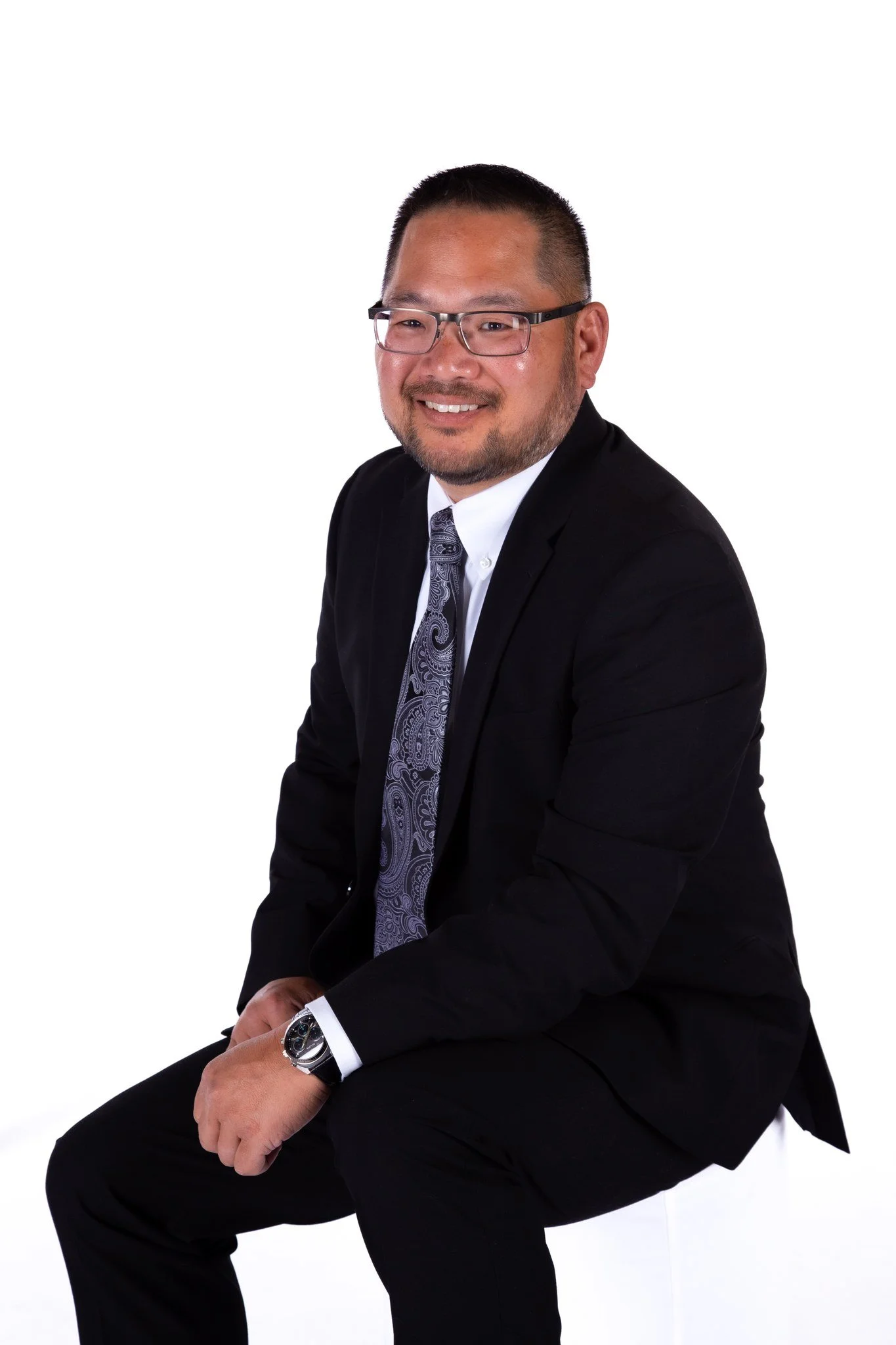 A man in a black suit, white shirt, and patterned tie sitting on a white stool against a white background.
