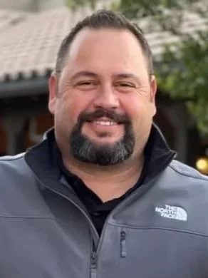 Close-up of a smiling man with dark hair and beard wearing a gray North Face jacket outdoors.