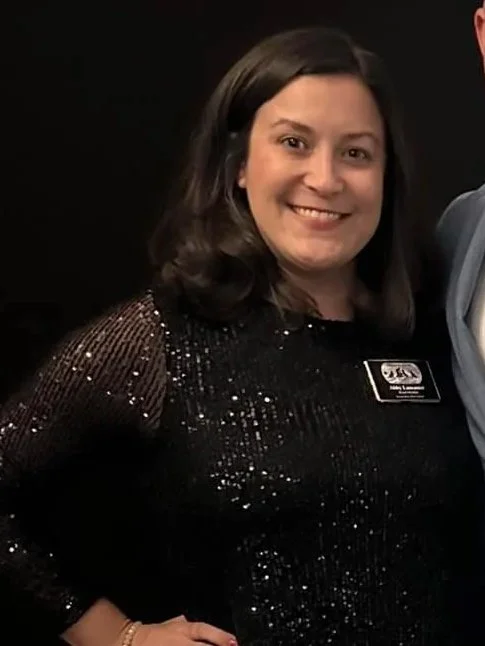 A woman with shoulder-length dark hair wearing a black, sparkly dress and a name tag, smiling at the camera.