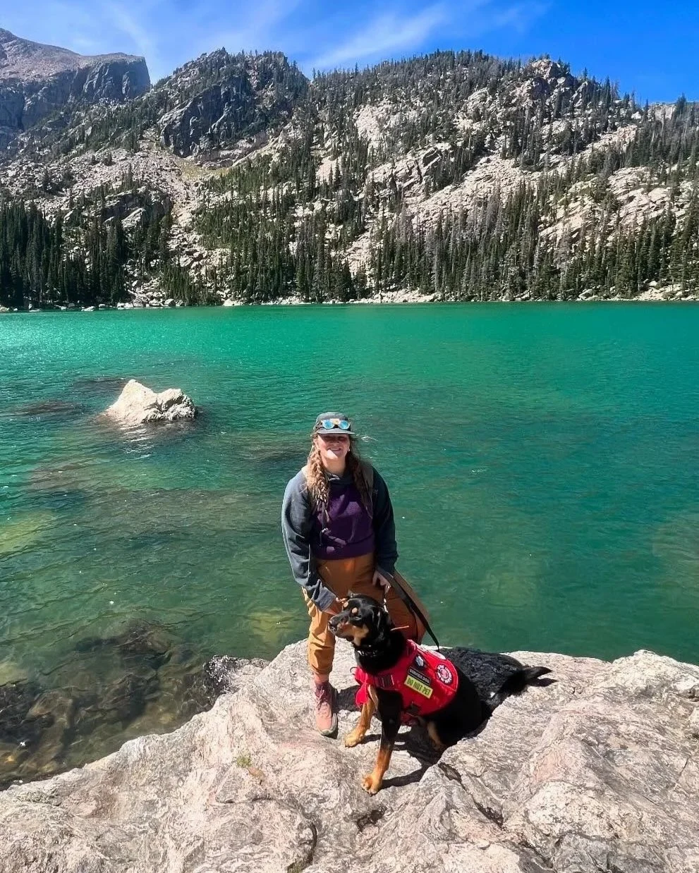A woman and a guide dog standing on rocks beside a turquoise lake with mountains and pine trees in the background, under a blue sky.