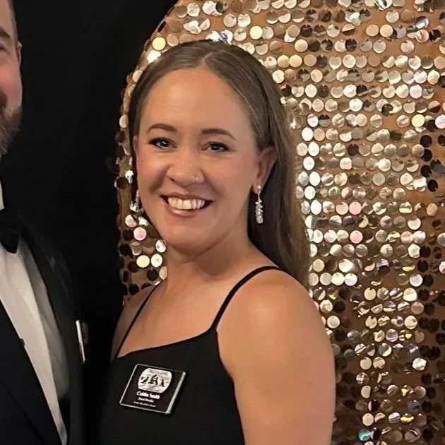 A smiling young woman with earrings wearing a black sleeveless dress and a name badge, standing in front of a sequined backdrop at a formal event.