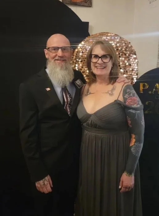 A man with a beard, glasses, and a suit, standing next to a woman with glasses, tattoos, and a strapless dress, both smiling at the camera, at an indoor event with a decorative glittery backdrop.