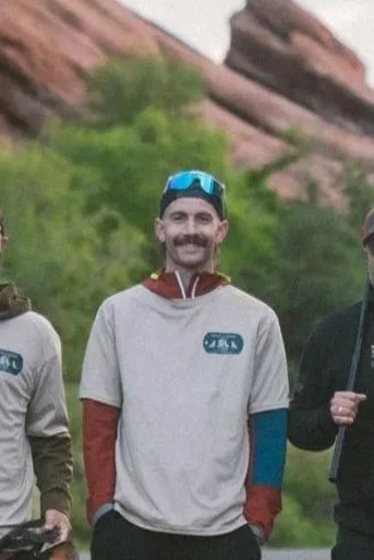 A man with a beard and mustache, wearing a beige athletic shirt with red and blue sleeves, sunglasses on his head, and standing outdoors with greenery and rocks in the background.