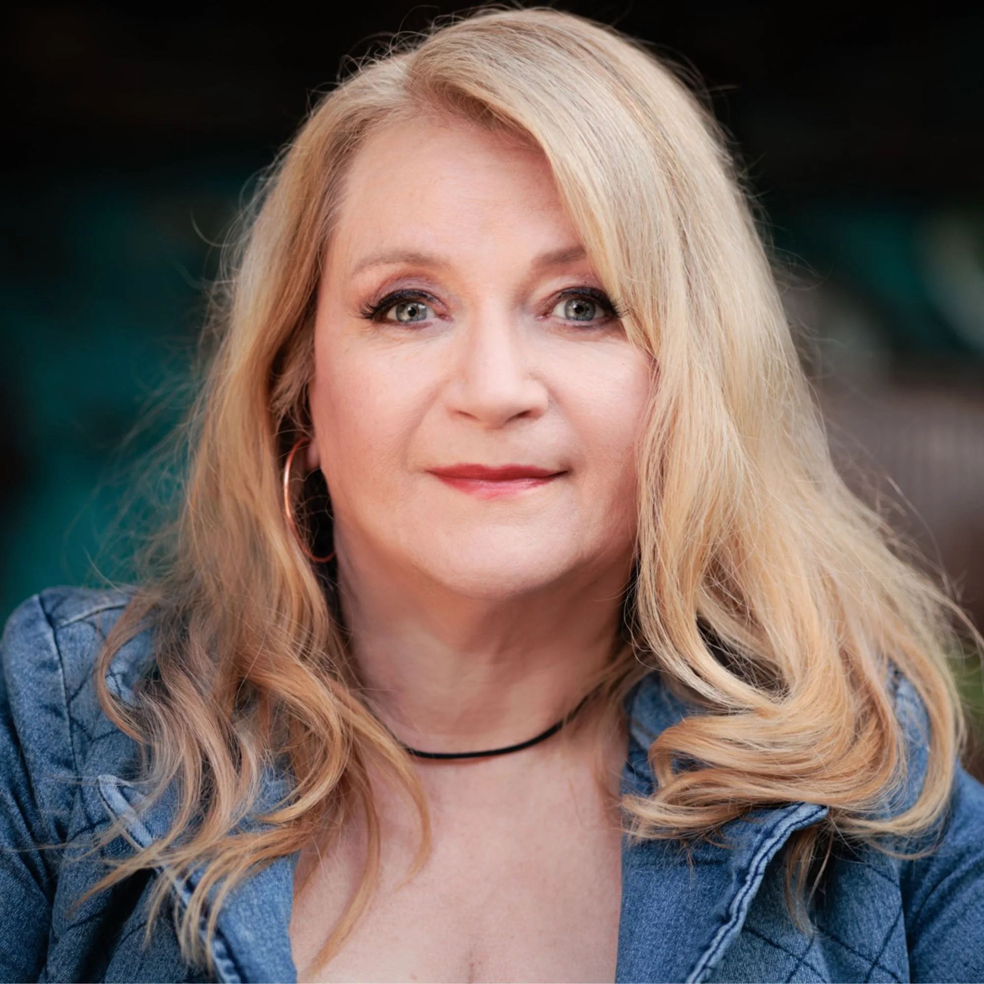 A woman with long blonde hair, wearing a denim jacket and hoop earrings, looking directly at the camera with a slight smile.