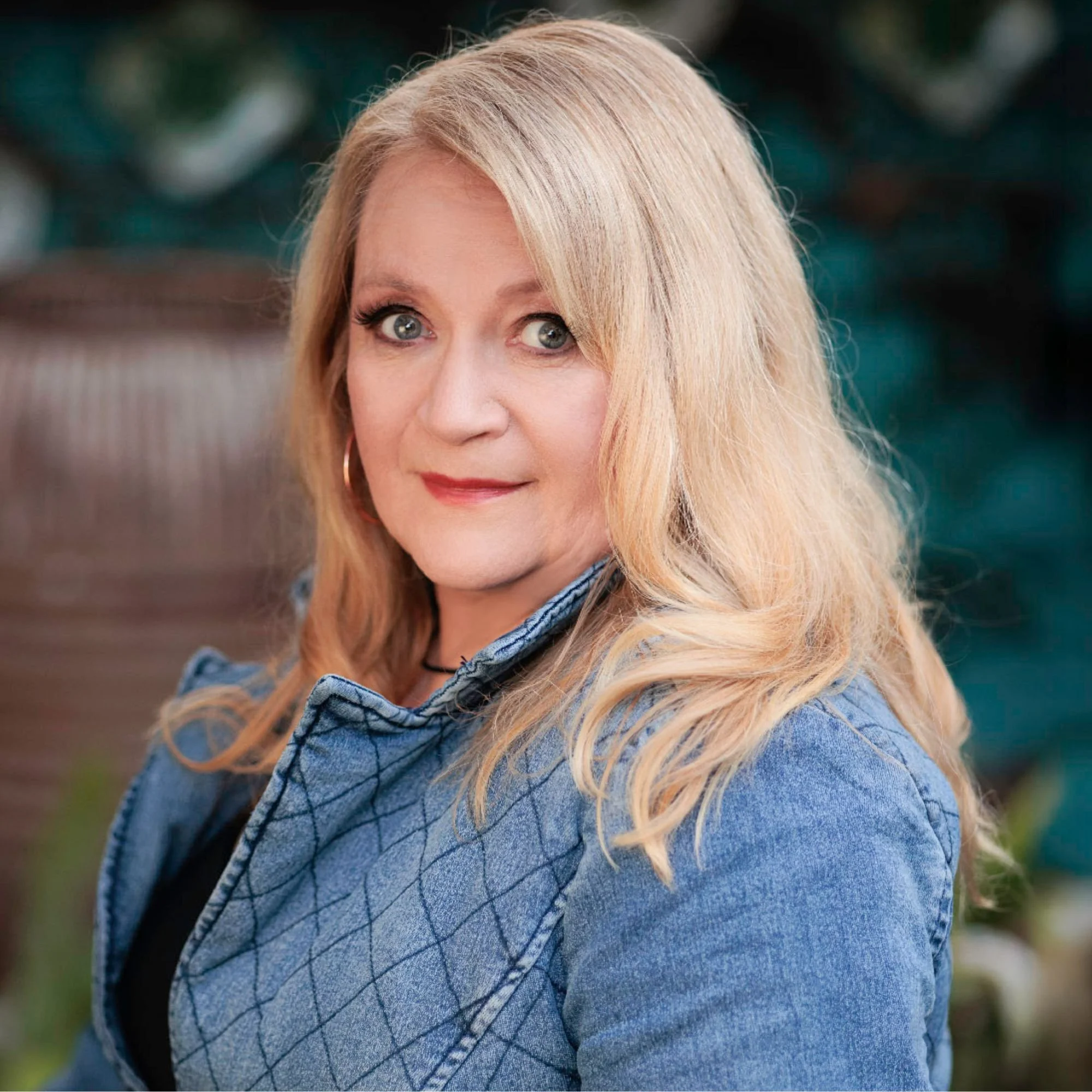 A woman with blonde hair, light skin, and blue eyes, wearing a denim jacket and a black top, posing outdoors with a blurred natural background.