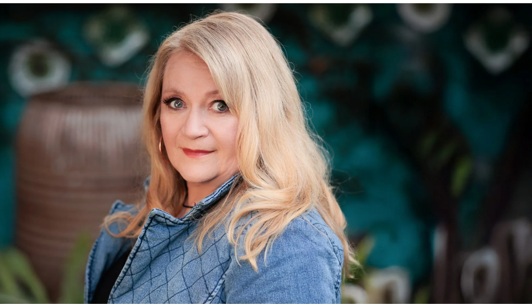 A blonde woman with blue eyes and hoop earrings, wearing a denim jacket, posing outdoors with greenery and a large ceramic pot in the background.