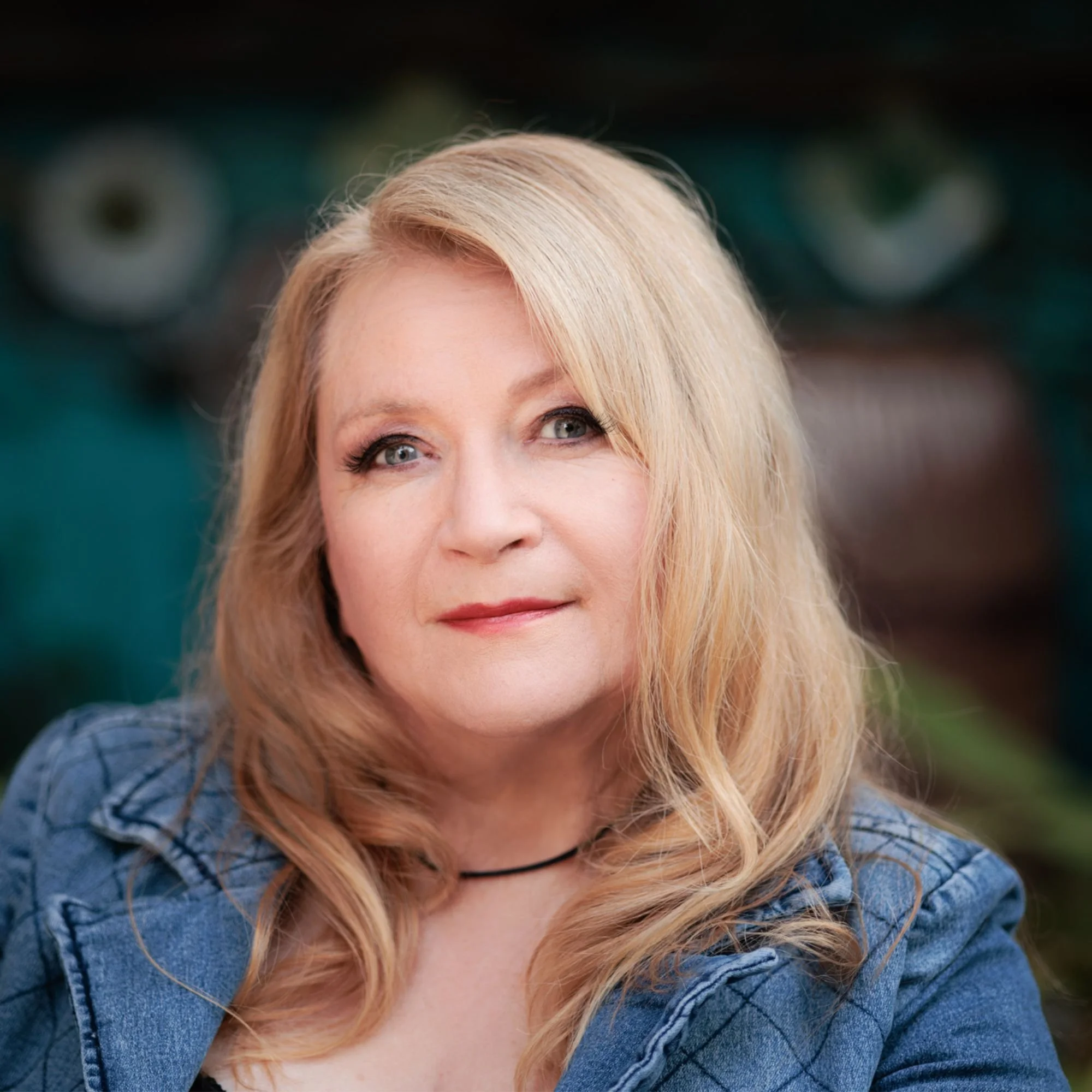 A portrait of a middle-aged woman with long blonde hair, wearing a denim jacket and a black choker necklace, smiling softly in front of a blurred background.