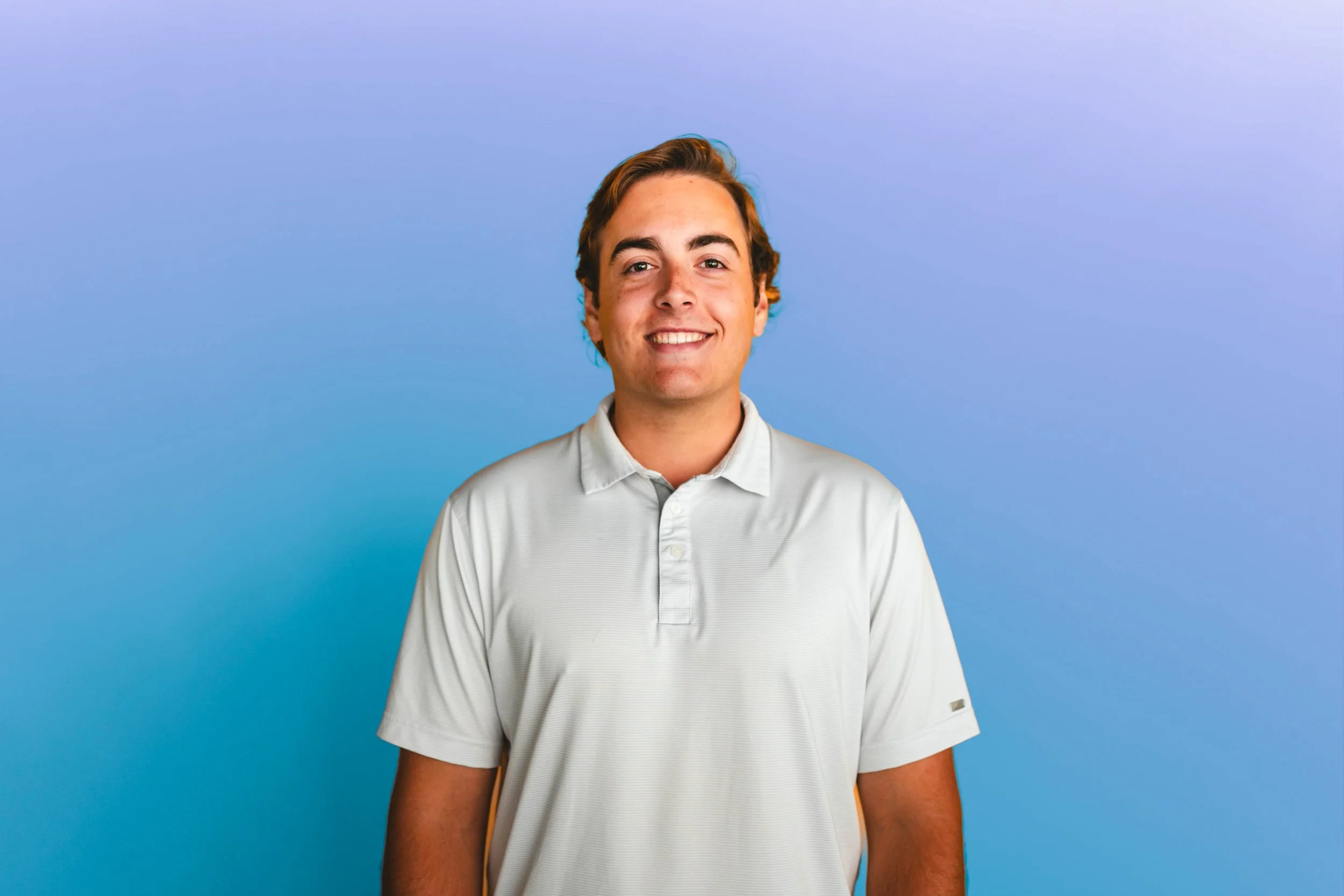A young man with light brown hair and a bright smile, wearing a white polo shirt, standing against a gradient blue background.
