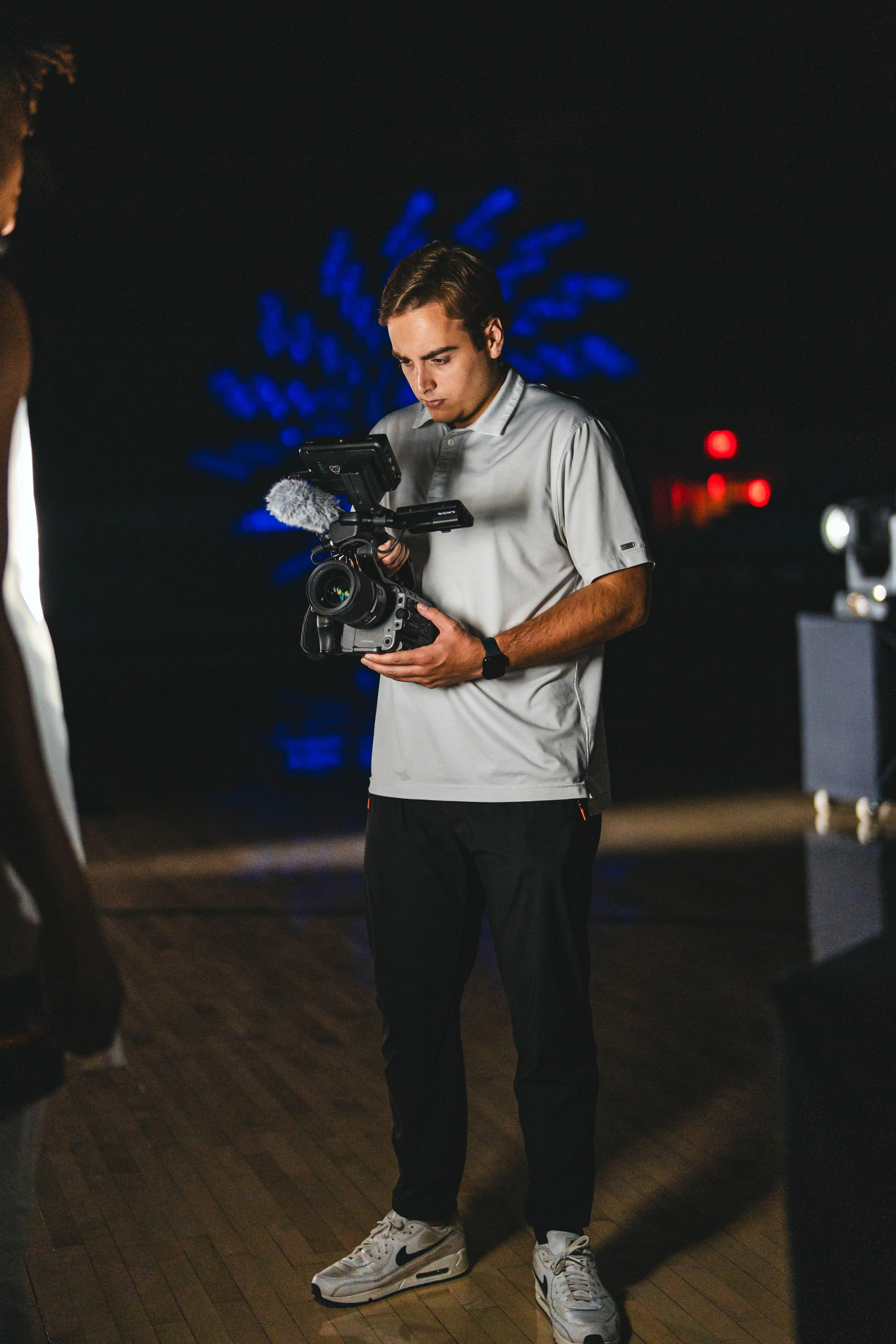 Young man operating a professional video camera at night with city lights and a blue-lit tree in the background.