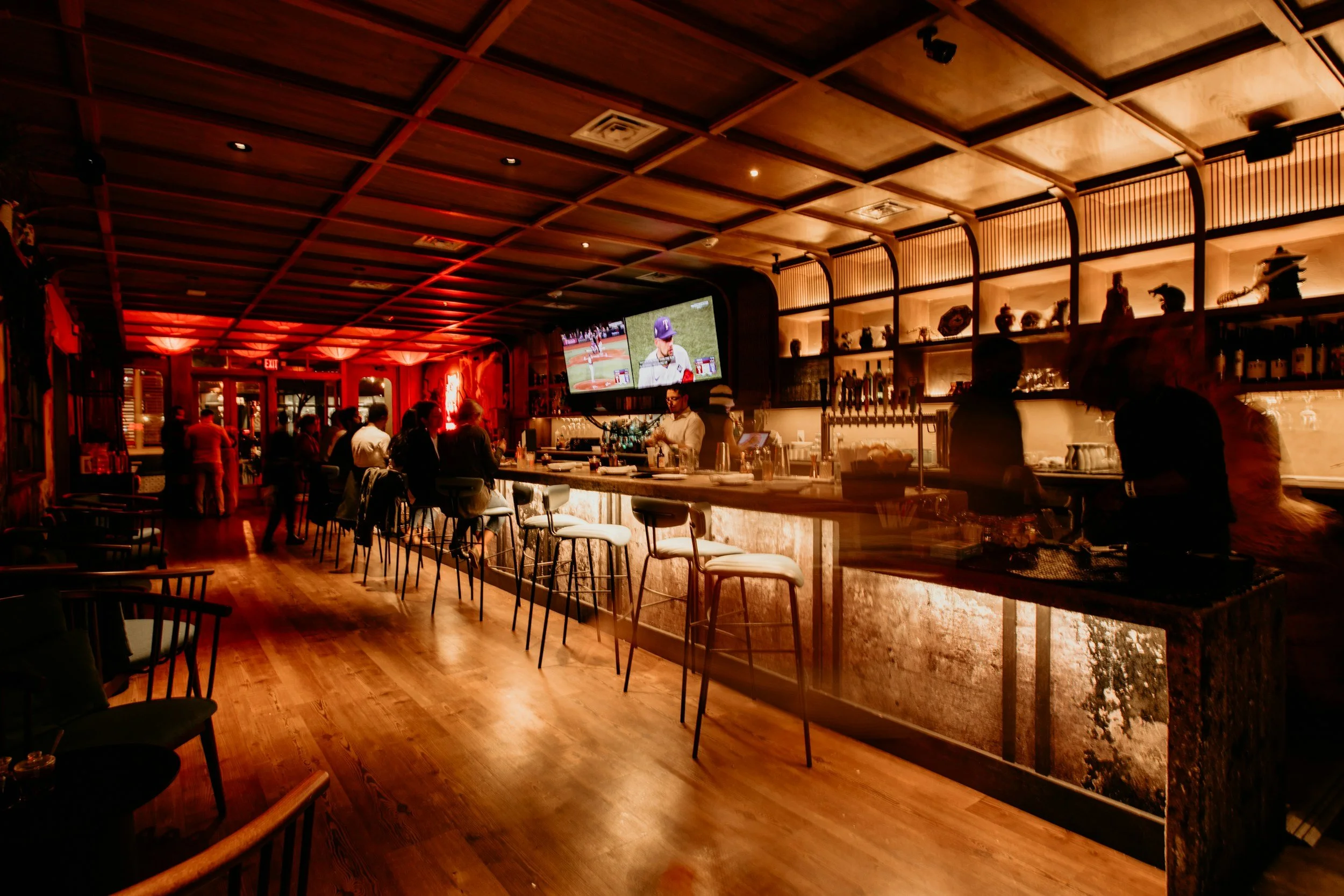 A lively bar and restaurant with warm lighting, a wooden ceiling, and a long bar counter. People are seated at the bar and standing around, enjoying drinks. A bartender is preparing drinks, and a large TV above the bar displays a baseball game.