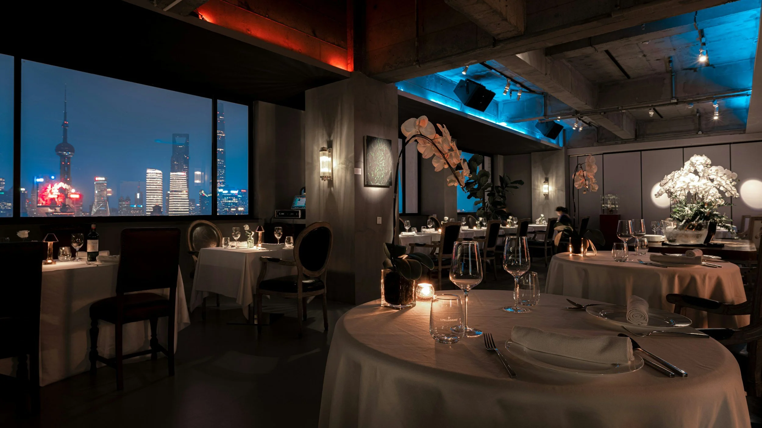 Candlelit fine dining room with white-linen tables overlooking a city skyline at night