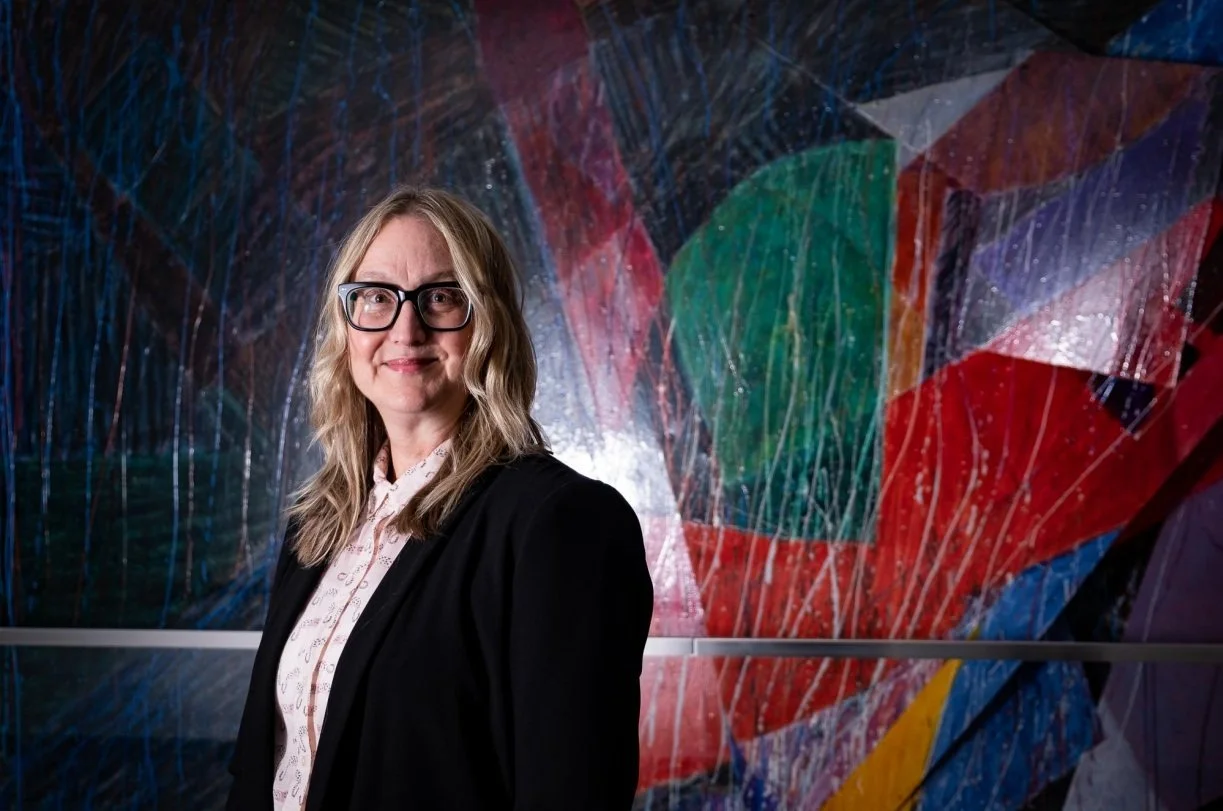 Portrait of Meg Shiffler, Director of AST, a white woman with blonde hair and glasses standing in front of a piece of artwork