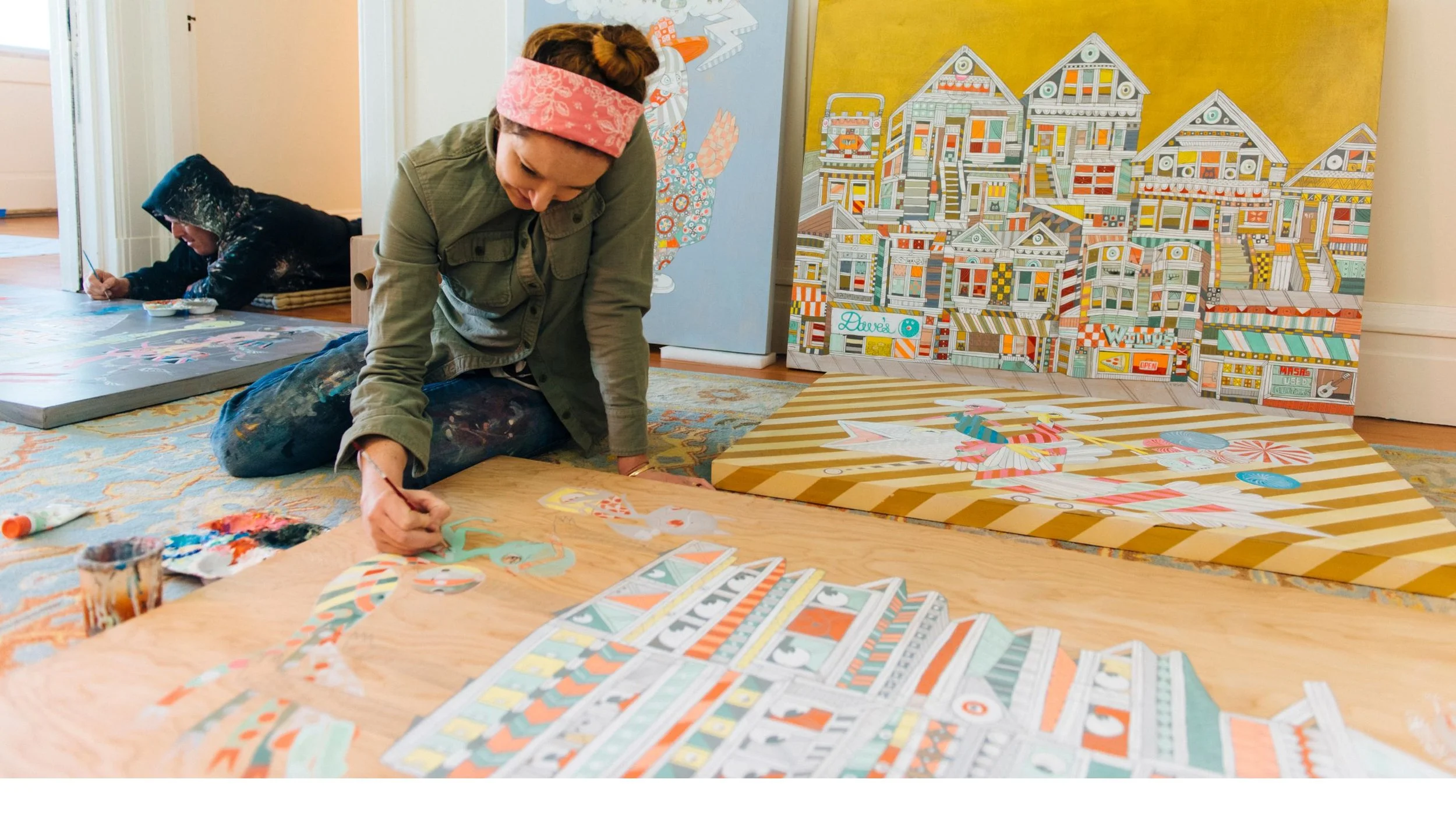 Two artists paint on canvases while sitting on the floor