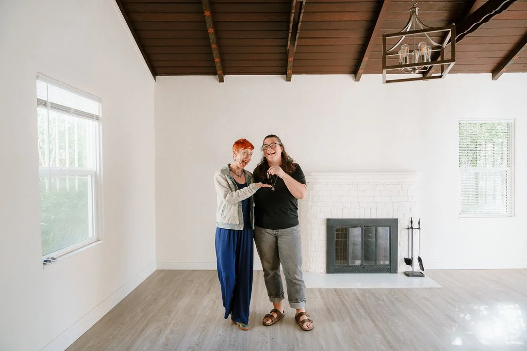 Two women stand together in an empty house, one holding new house keys and one showing them off
