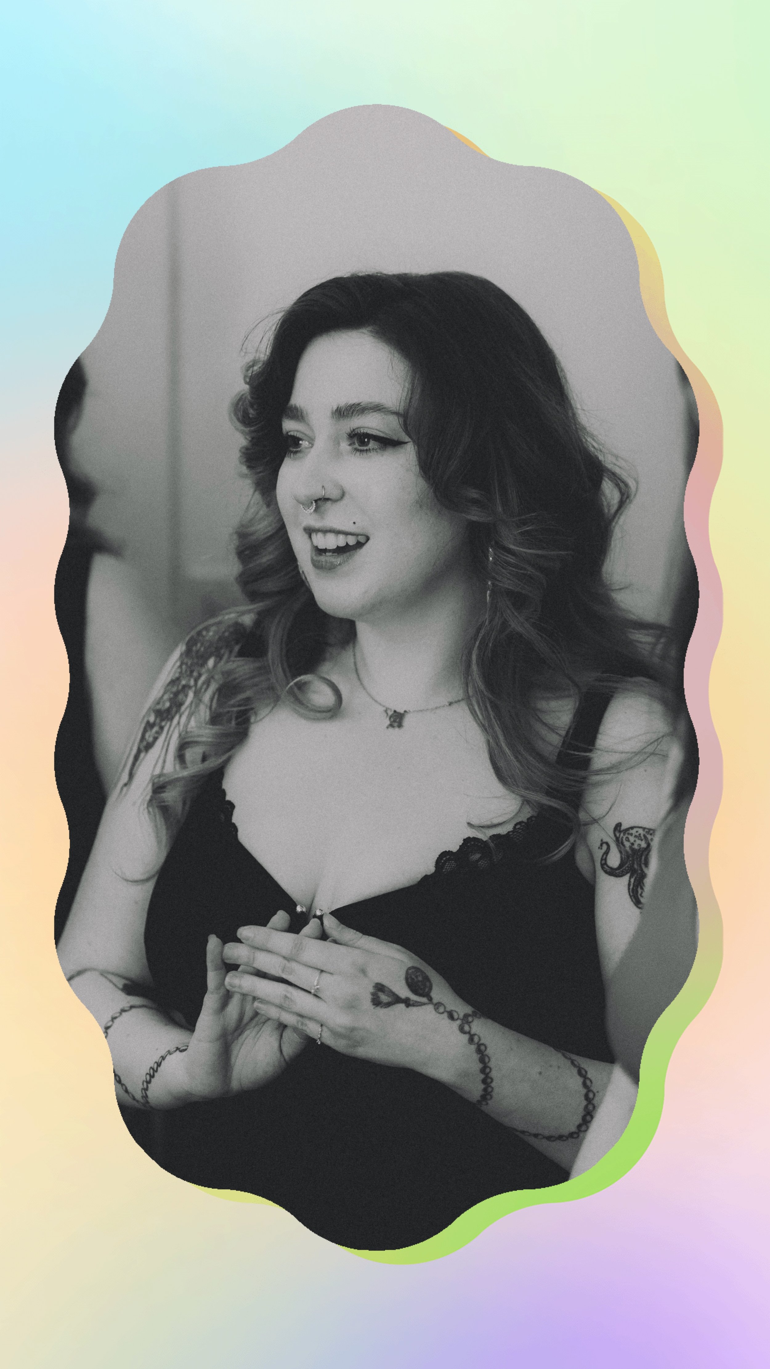 A woman with dark, wavy hair, tattoos on her arms and shoulder, wearing a black top, smiling, and looking to her left. She has a septum piercing, a nose piercing, and is wearing a necklace and rings.