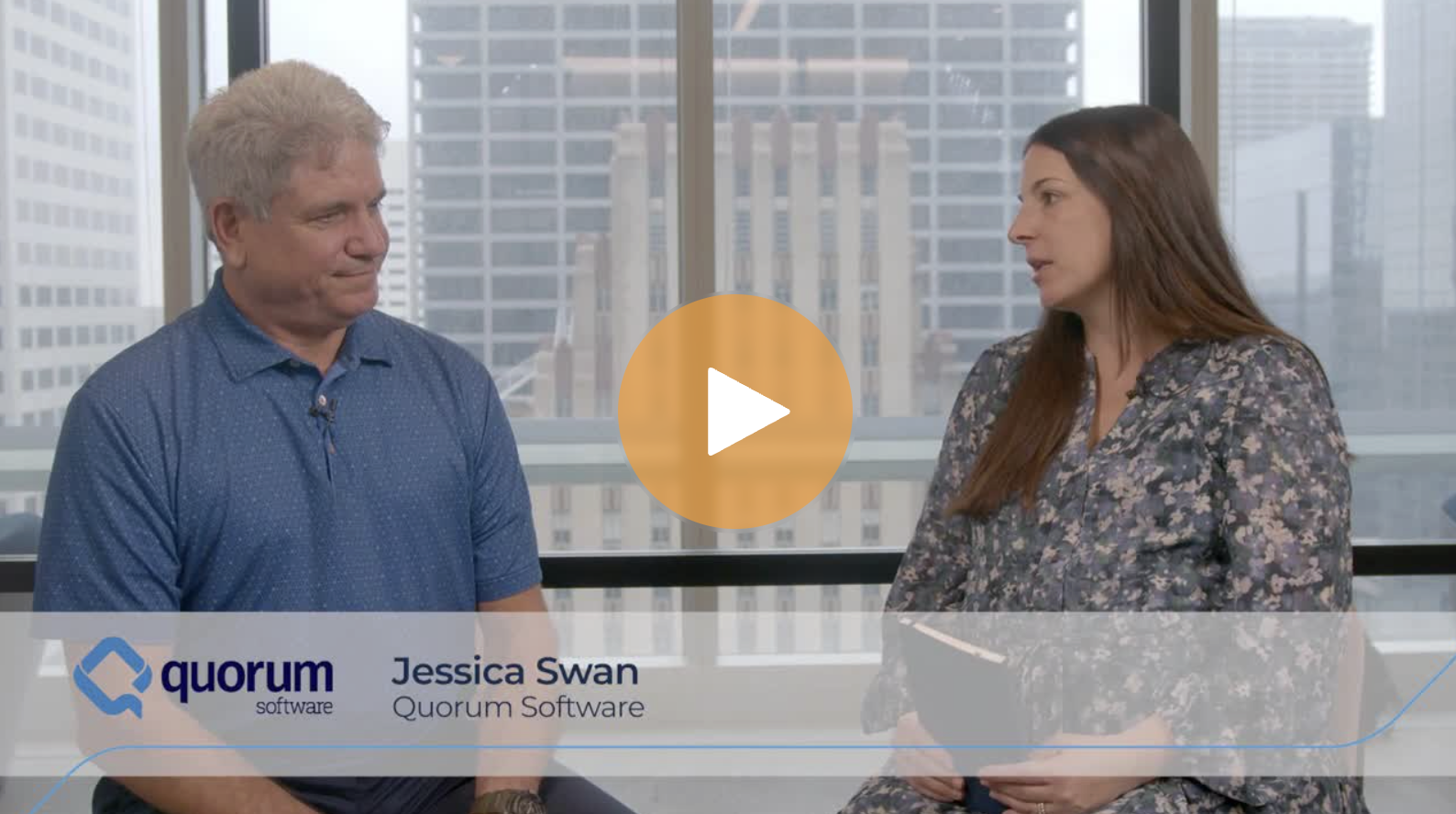 A man and a woman seated and talking in an office with large windows and city skyscrapers in the background. The man is wearing a blue polo shirt and the woman a floral blouse. A translucent banner at the bottom displays the Quorum Software logo, and the names Jessica Swan and Quorum Software.