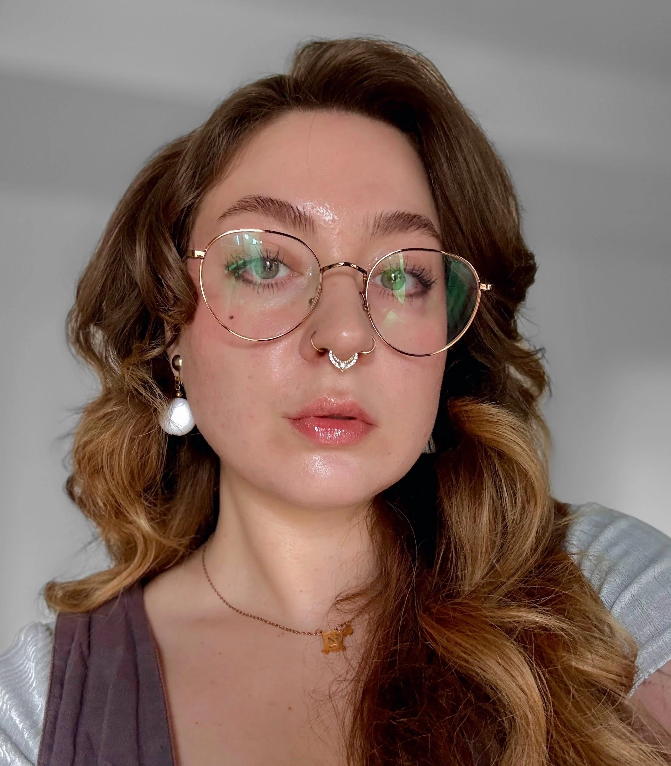 A woman with wavy red hair, wearing round glasses, a septum ring, a gold necklace, and earrings, looking directly at the camera against a plain background.
